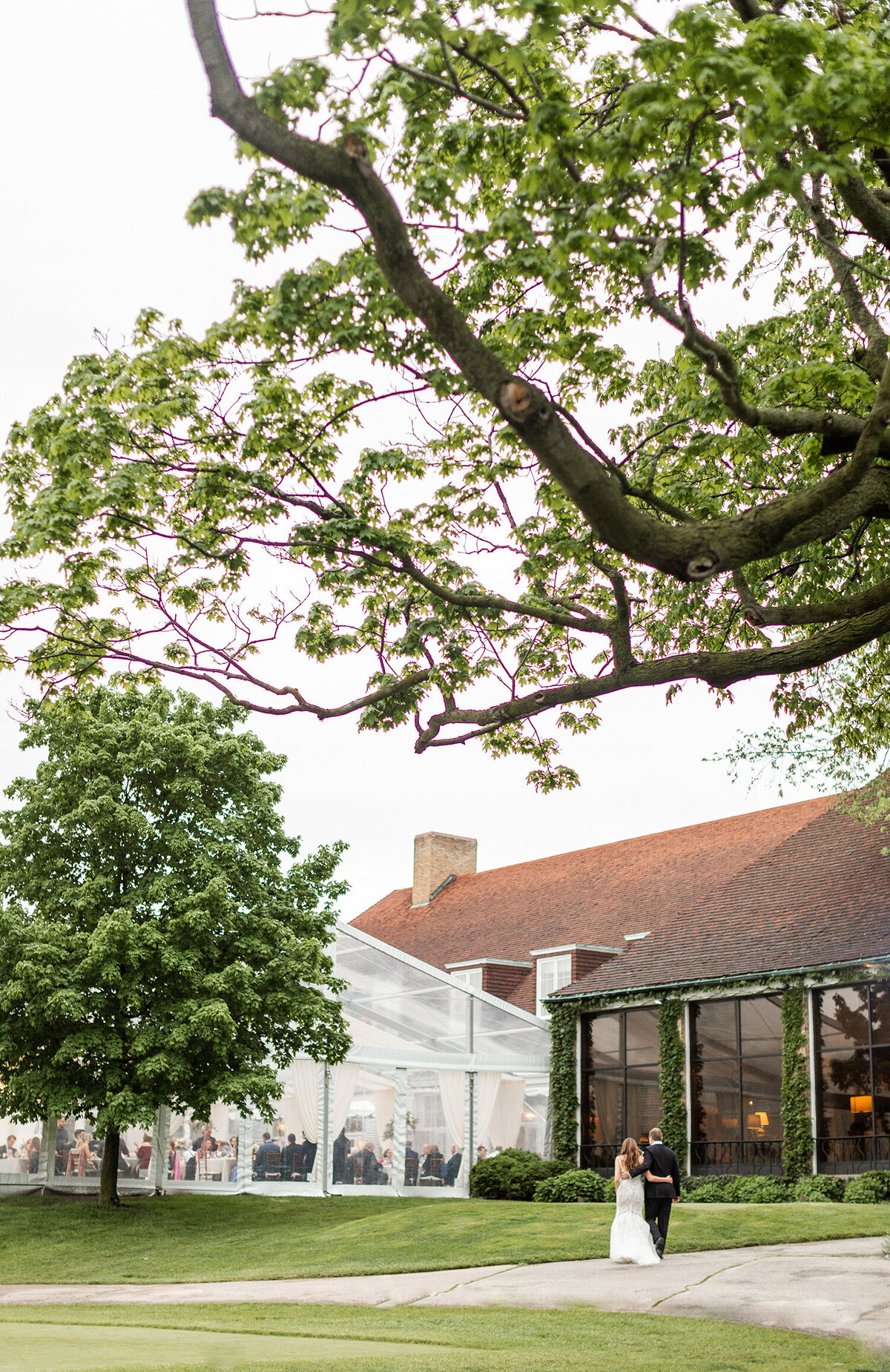 Outdoor wedding venue with a clear tent and greenery, couple walking away together