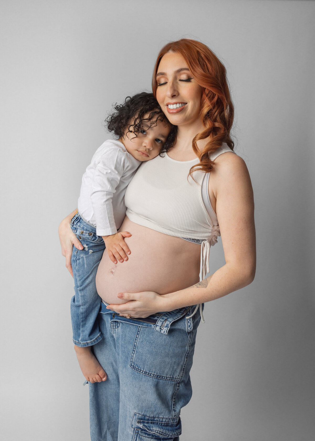 A sweet family moment as a young child embraces a pregnant mother wearing a white crop top and jeans against a gray backdrop in NJ.