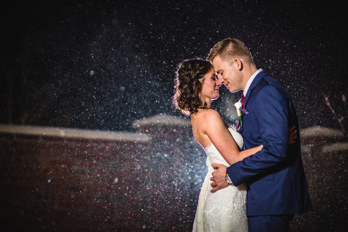A Columbus wedding photographer captures a magical moment of a bride and groom embracing under a flurry of snowflakes at night. The groom, dressed in a navy blue suit, holds the bride close, who stuns in a strapless lace gown. Their faces touch tenderly, illuminated by the light that also highlights the snow, creating a sparkling, enchanting atmosphere around them.
