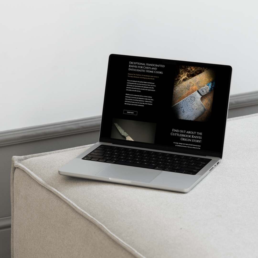 A laptop displaying a website for Cuttlebrook Knives, placed on a light grey upholstered surface. The screen shows an image of an intricately designed chef knife with accompanying text promoting handcrafted knives for chefs and enthusiastic home cooks. The page also features a section inviting users to learn about the origin story of Cuttlebrook Knives. The background is simple and minimalistic, focusing attention on the laptop screen.