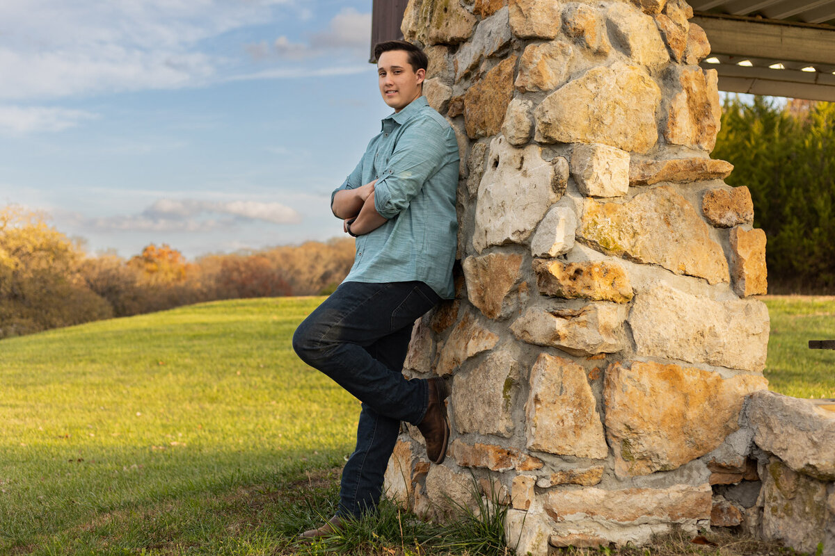 A senior boy leaning up against a stone wall with his foot resting up against of it in Lawrence, KS