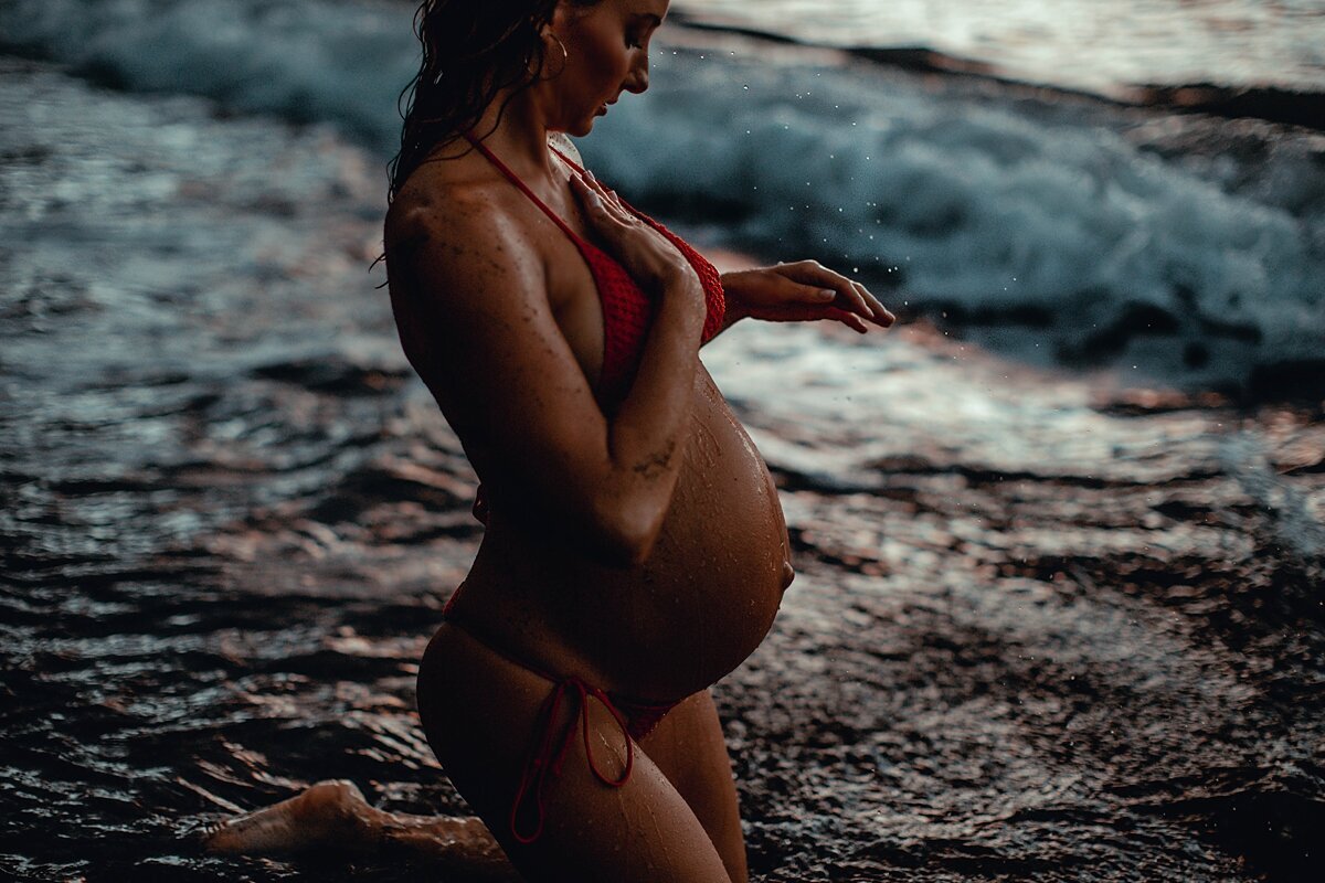 Candid maternity portrait of a woman wearing a red bikini wiping water from her chest on the beach in Wailea.