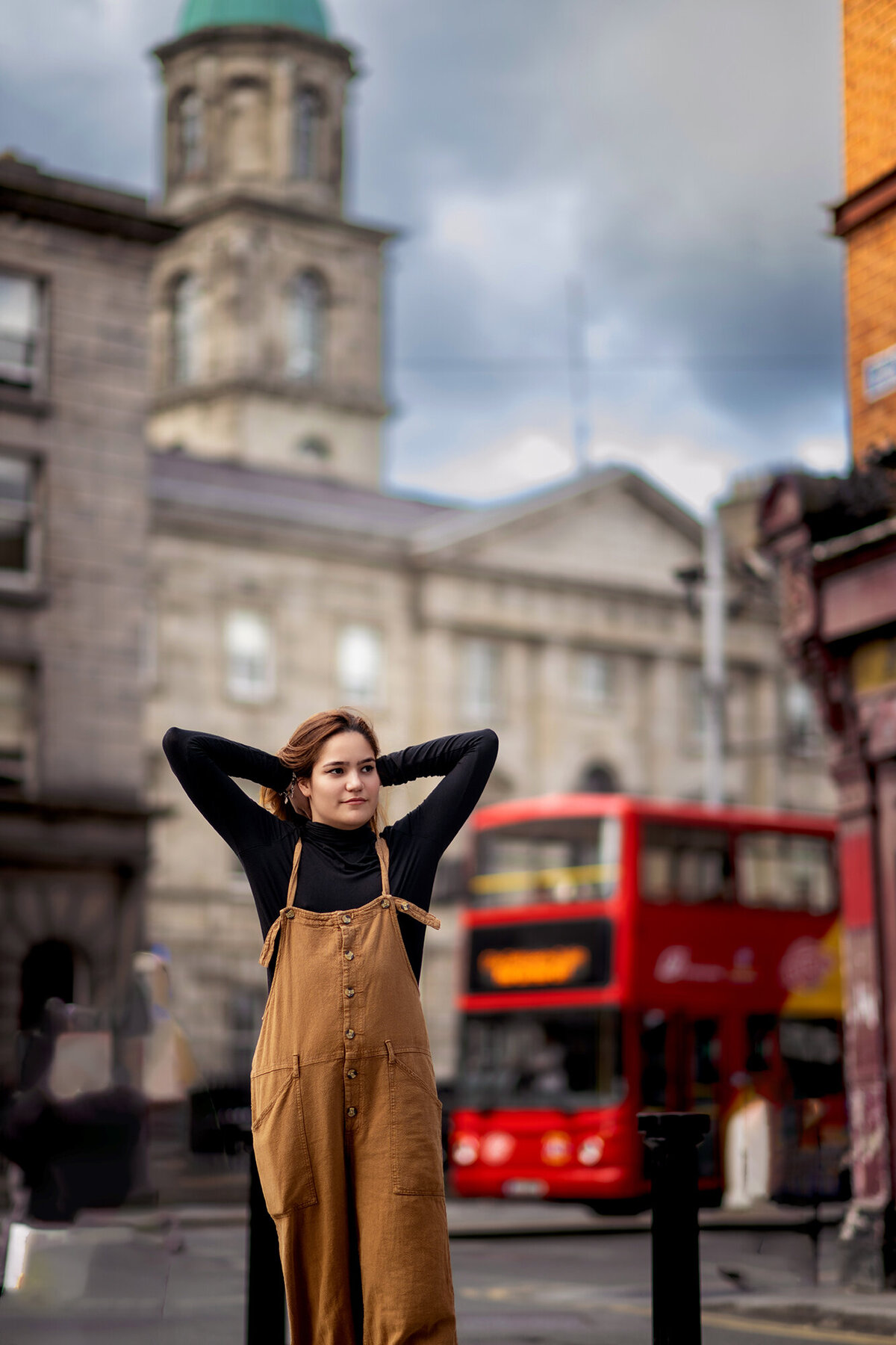 A high school senior stands in a London street with hands behind her head in brown overalls and black top