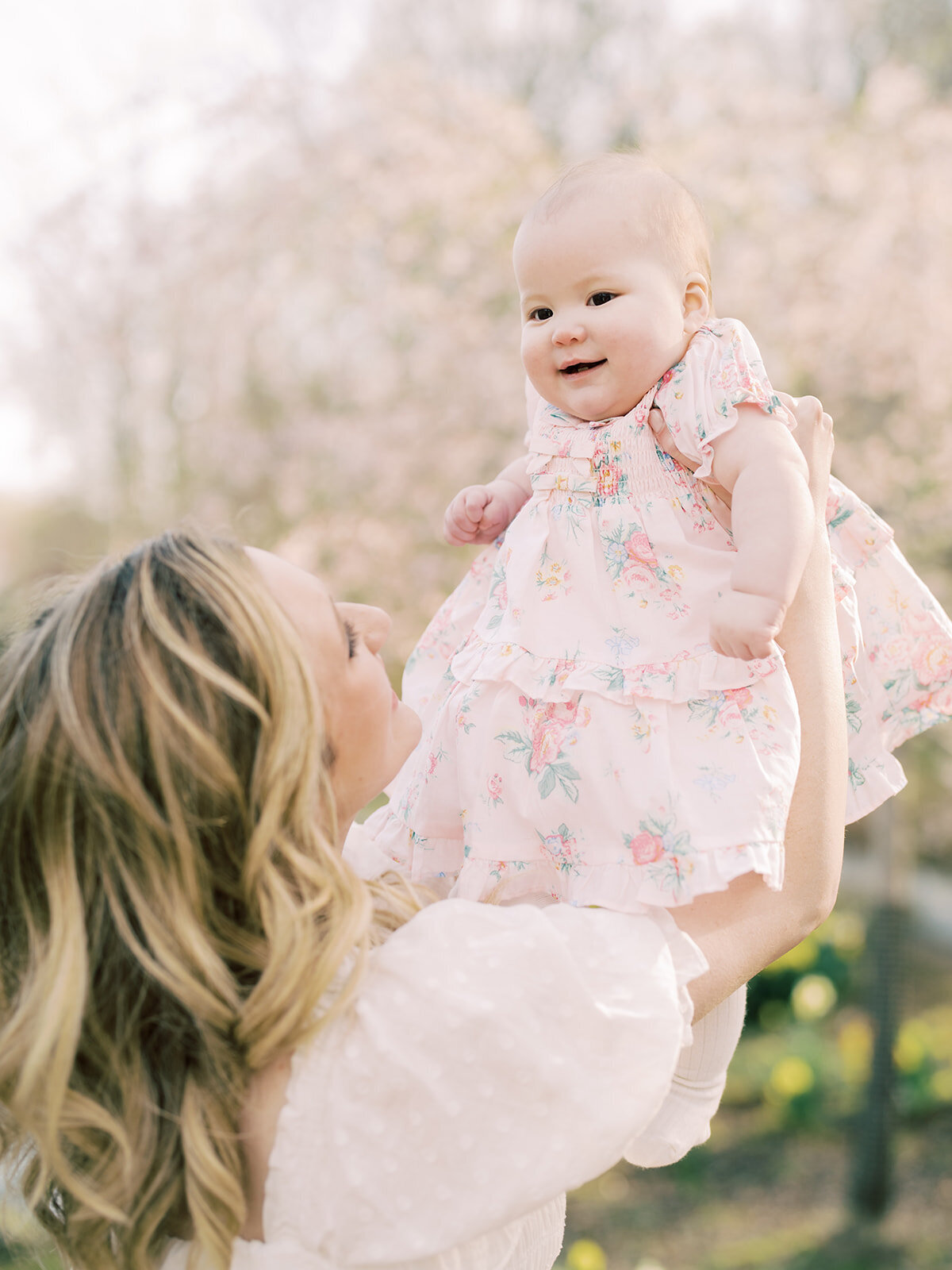 Blonde mother holds baby girl up in the air near cherry blossom tree.