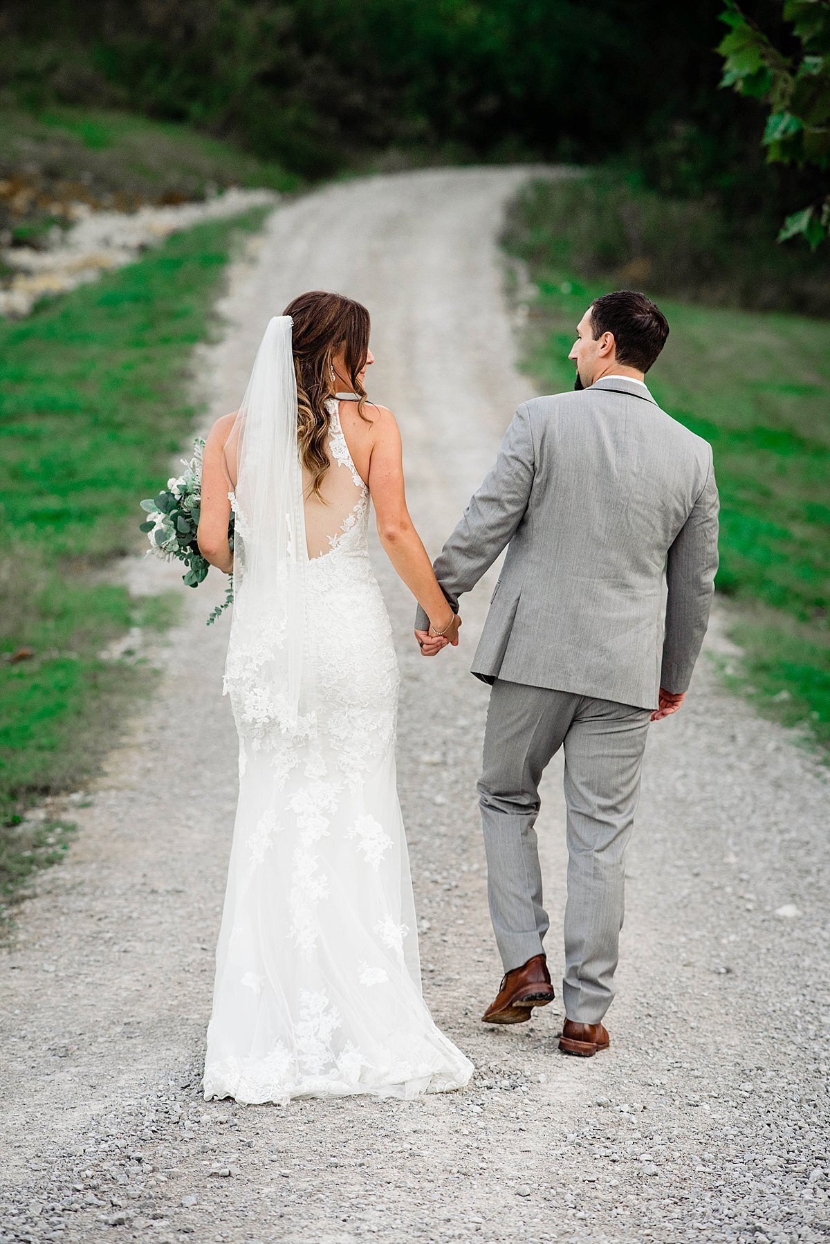 The bride and groom walk away from the camera down a gravel path. with greenery and tress on either side. The  groom is wearing a light gray suit with brown shoes.  The bride is wearing a fitted lace wedding dress with an illusion back and a sweep train. She has a fingertip length veil and a bouquet of greenery and white flowers. They are holding hands as they walk away.