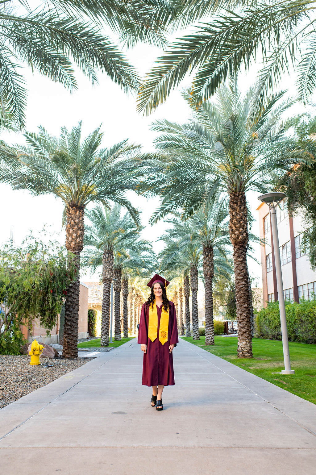 A graduate in a cap and gown walking down a path surrounded by palm trees.