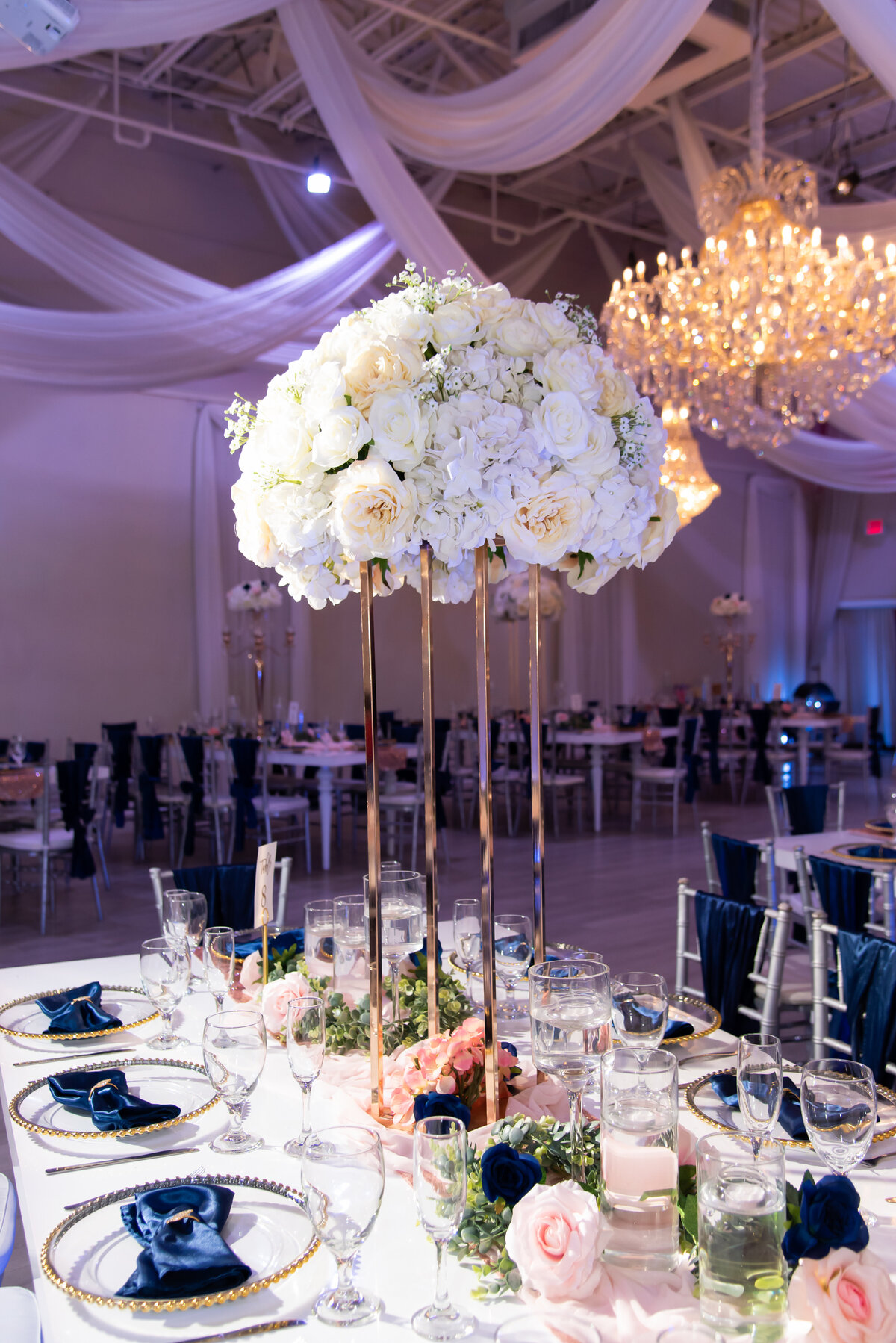 navy and blush design reception with tall gold centerpiece pillars and ceiling drapery with purple uplighting and crystal chandeliers 