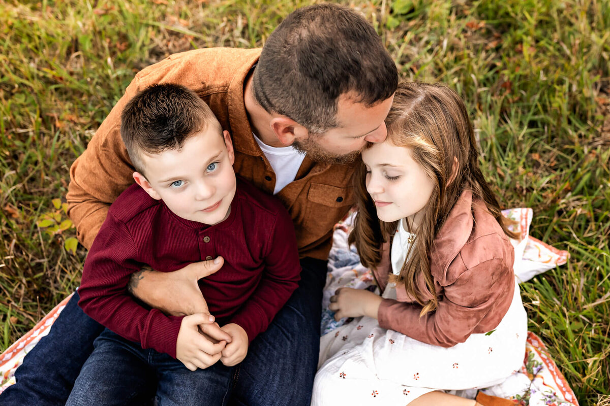 A man sits on a blanket in a grassy area, embracing two children. Capturing this heartfelt outdoor portrait, he kisses the head of a girl with long hair while the boy with short hair gazes at the camera. Everyone appears relaxed and content in this moment cherished by their Bloomington family photographer.