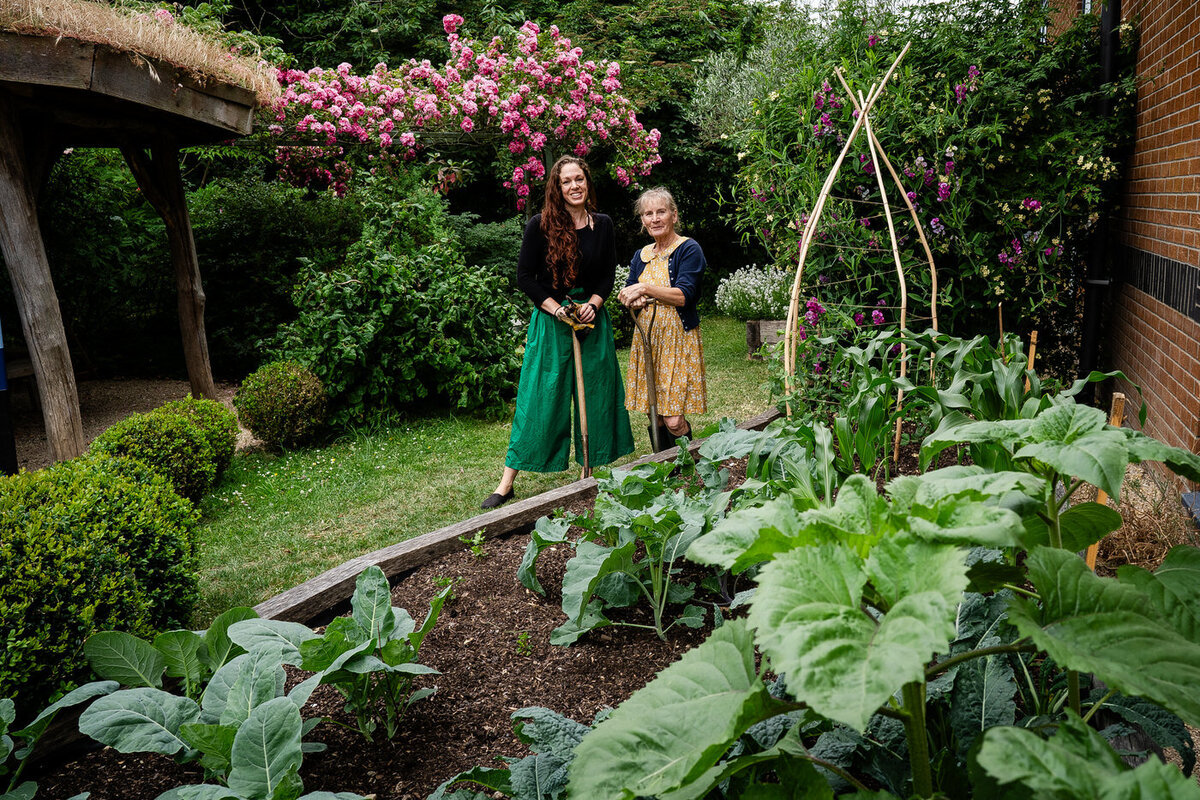 press release photograph of two women by a vegetable patch holding pitch fork and spade.