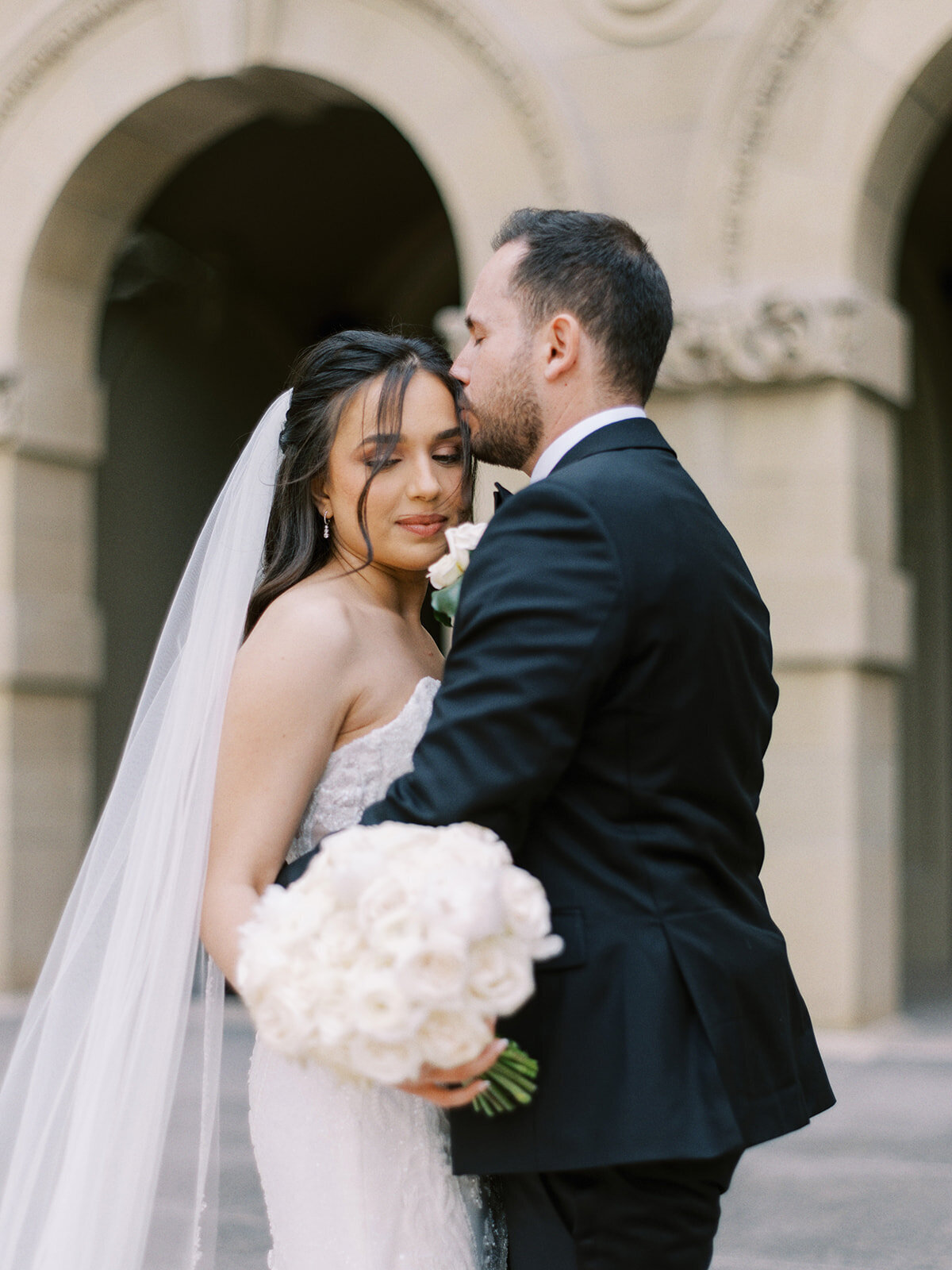 A bride holding a white bouquet stands next to a groom in formal wear who is kissing her forehead. They are in front of an archway at a classic Fairmont Palliser wedding in Calgary.