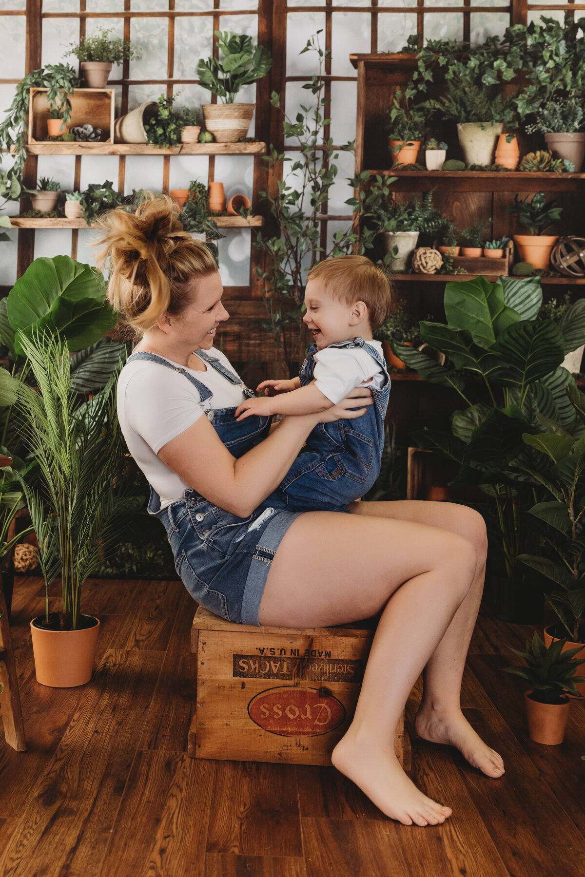 mom and toddler laugh together on a crate in a greenhouse