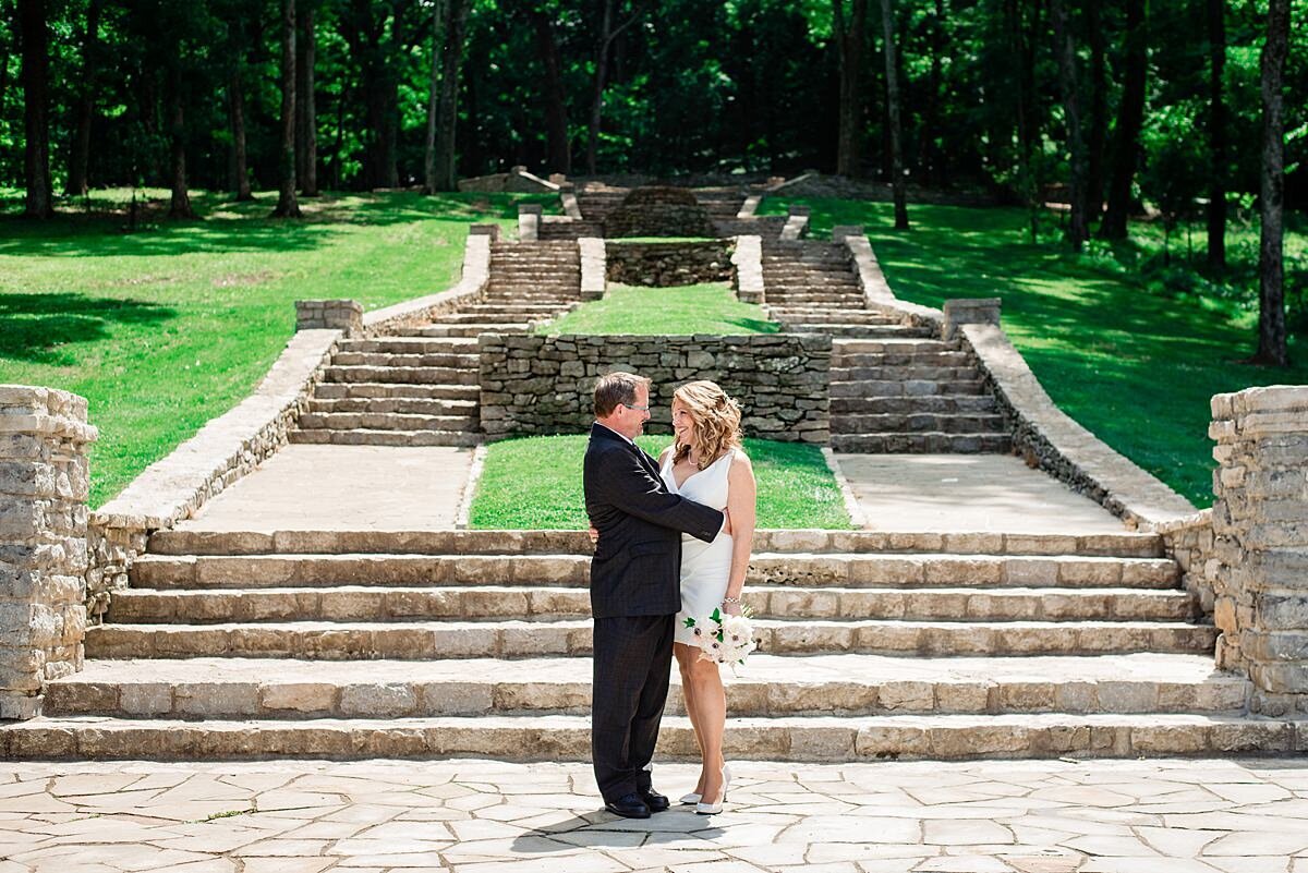 At the base of a long set of stone stairs that go in multiple levels , the groom embraces the bride. The groom is wearing a charcoal gray suit and the bride is wearing a short fitted white dress with a plunging neckline and an asymmetrical hemline.