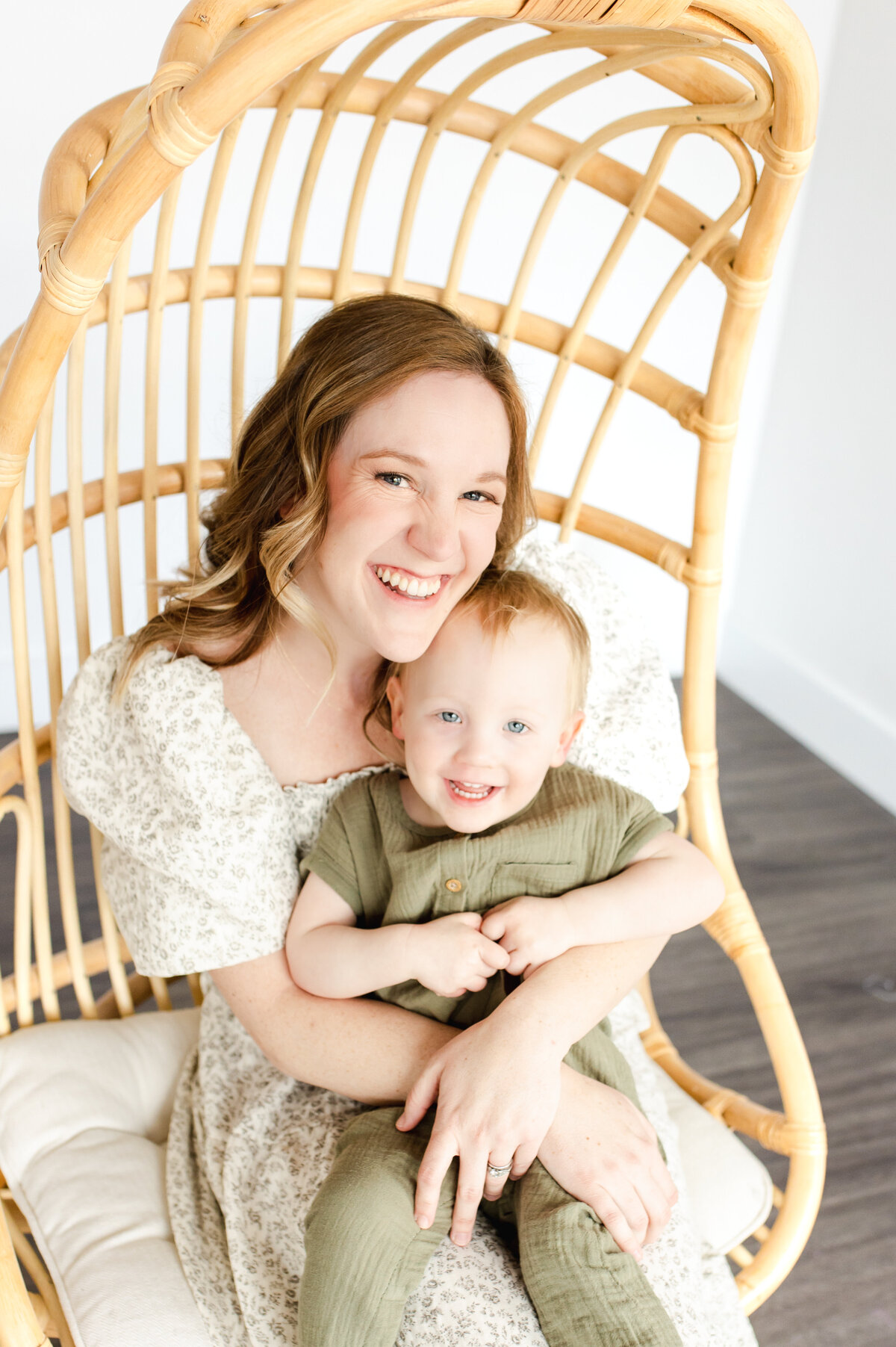 A brown haired woman sits in a rattan chair wearing a floral dress holding a toddler boy wearing a green outfit and sitting in front of a white backdrop