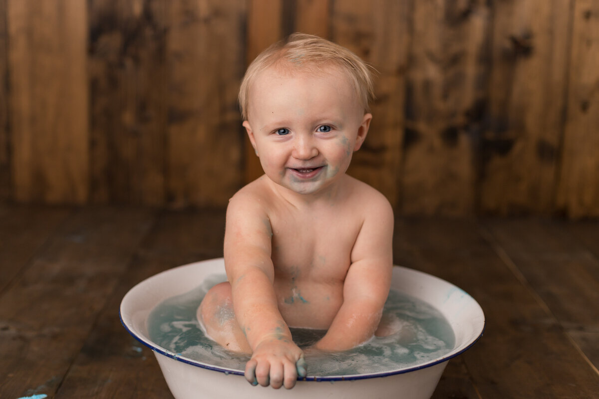 Baby sitting in the tub smiling, enjoying bath time after the cake smash, with remnants of blue frosting around.