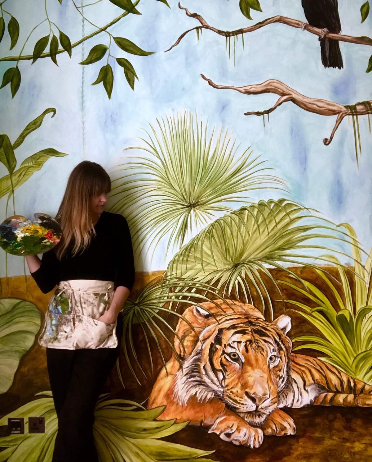 Brandy Klindworth of art and design studio Ladder Mouse mural of Lake City Minnesota stands in front of jungle rainforest nursery children's bedroom mural feautring a hand-painted tiger and toucan and jungle foliage located in Dubai, United Arab Emirates, traveling artist muralist holding paint palette.