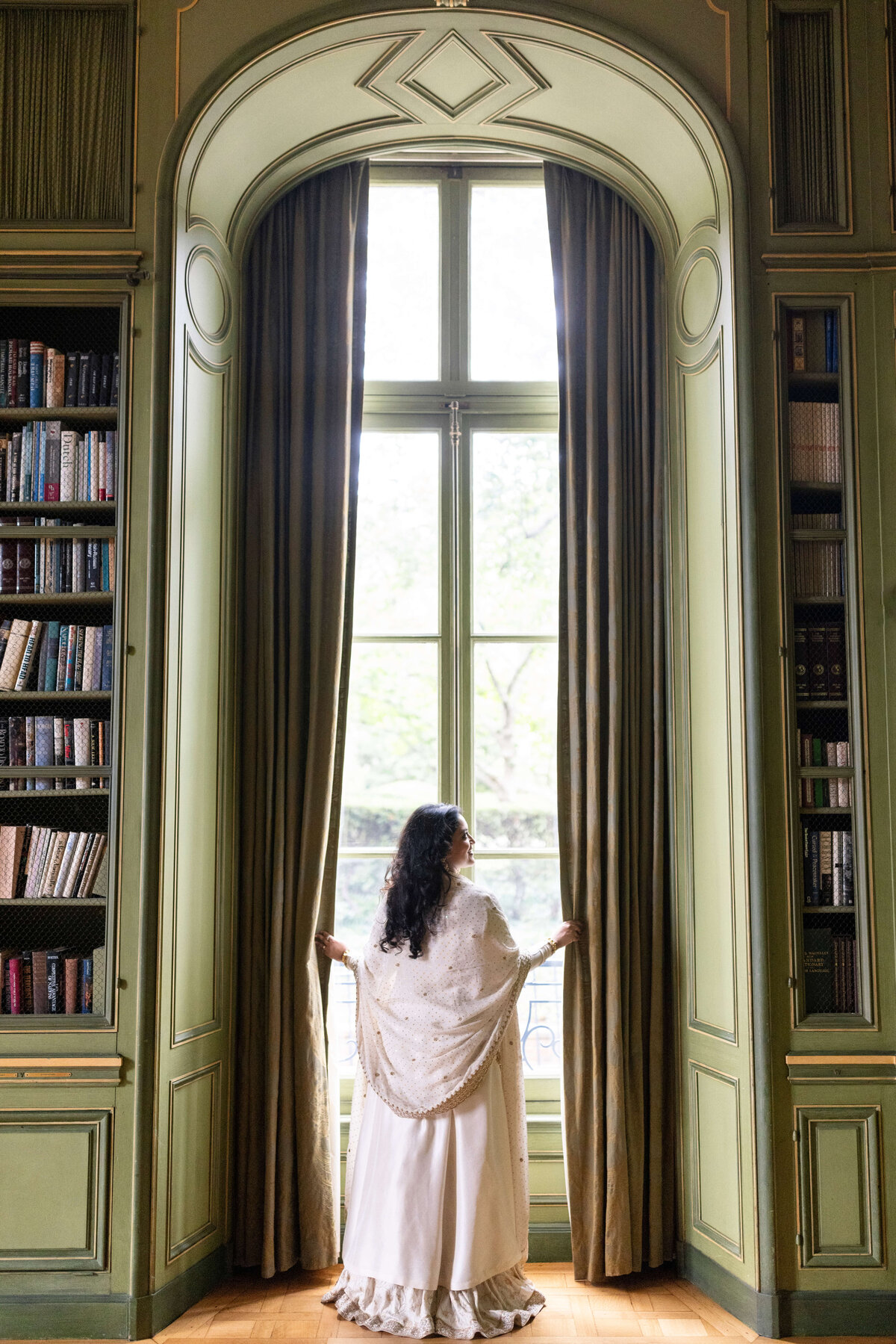 A woman in a white dress stands facing large, elegant windows framed by tall curtains in a library. Bookshelves filled with books are on either side of the window. The room appears ornate and historic.