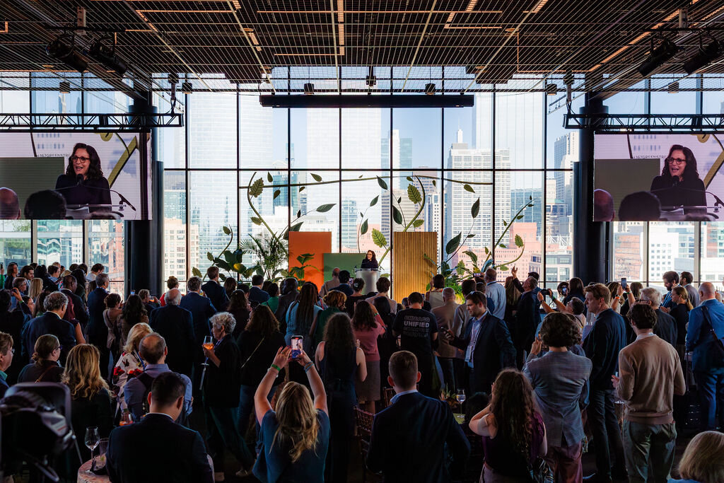 People stand around in an auditorium watching someone speak on stage.