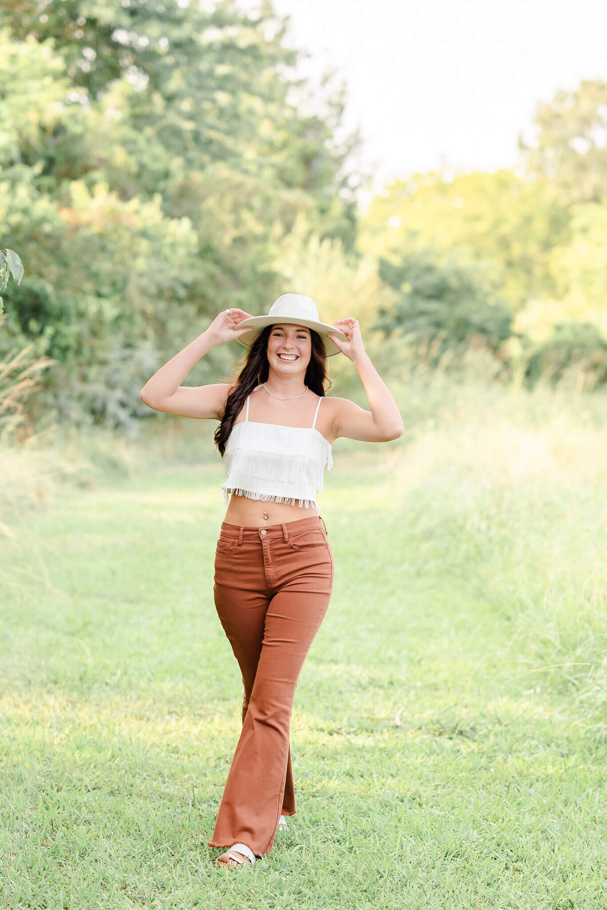 A high school senior, wearing orange pants and a white fringe top, holds her white hat and walks in a grassy field.