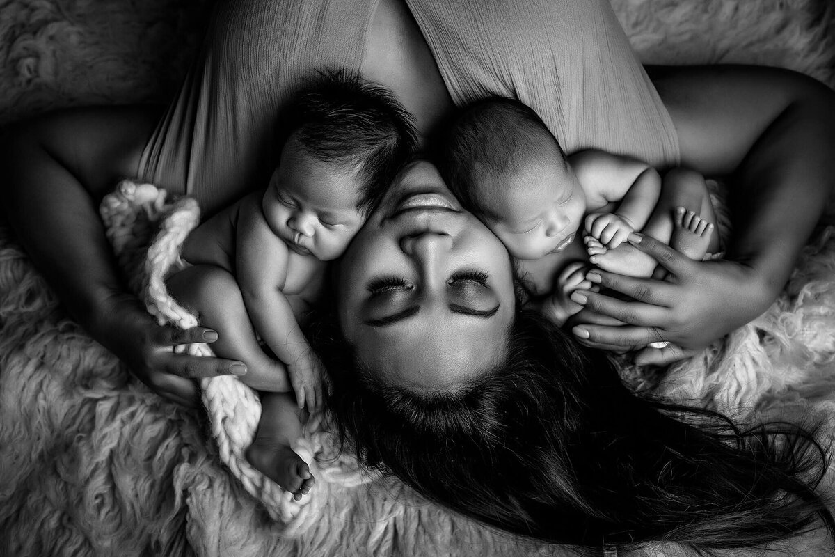 A woman lies on her back with her eyes closed, cradling two newborn babies on her chest. The babies sleep peacefully in this Orlando in-home newborn session. The black and white image creates a serene and tender atmosphere, showcasing the best of Orlando newborn photography.