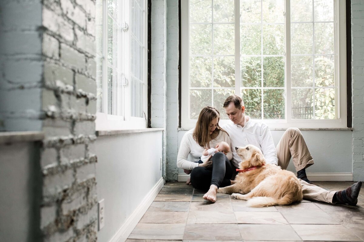 A couple and their dog sitting by a large window in a room with light-colored walls during an in-home newborn photography session.