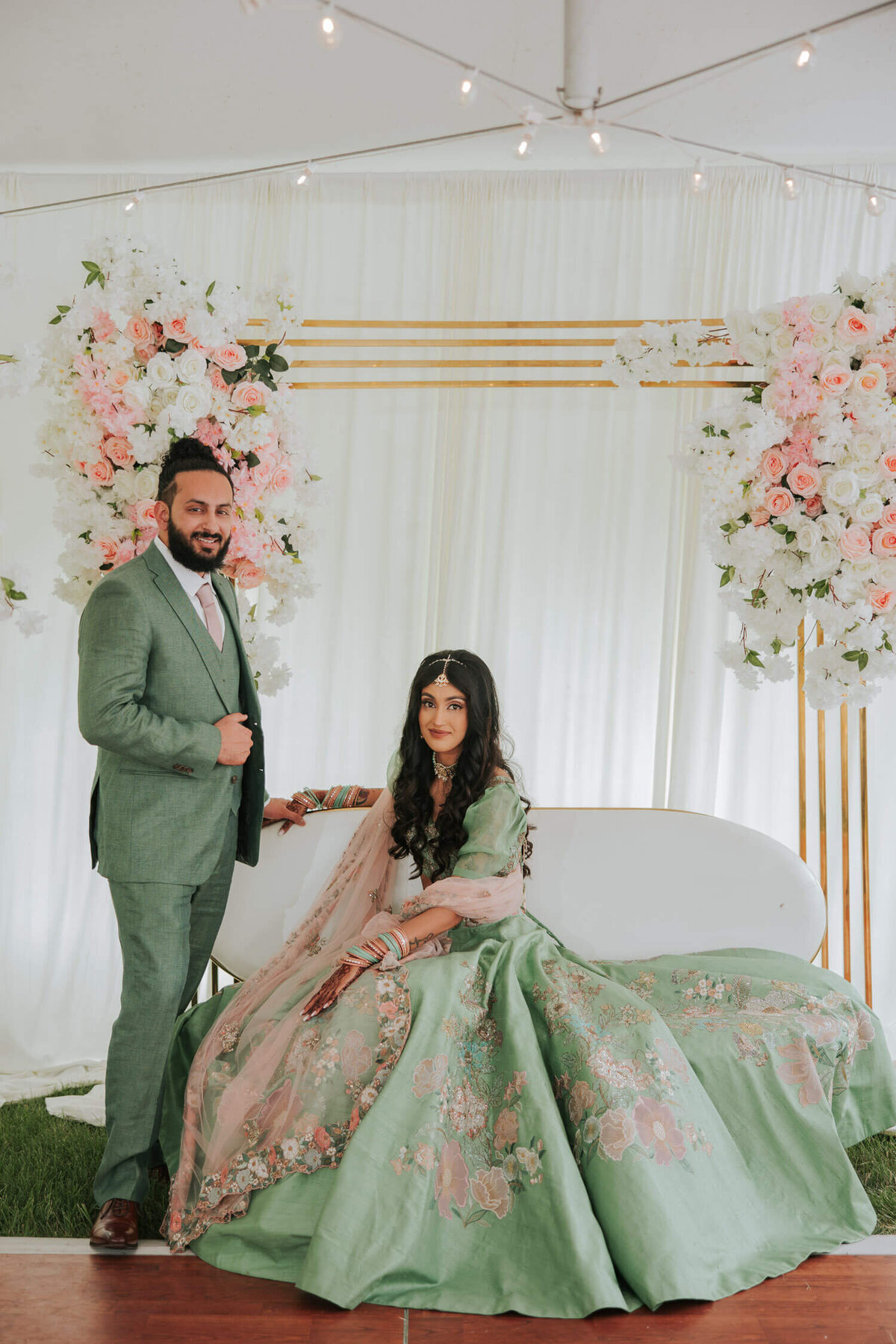 A couple poses in front of a floral backdrop, the bride seated gracefully in her intricate green attire