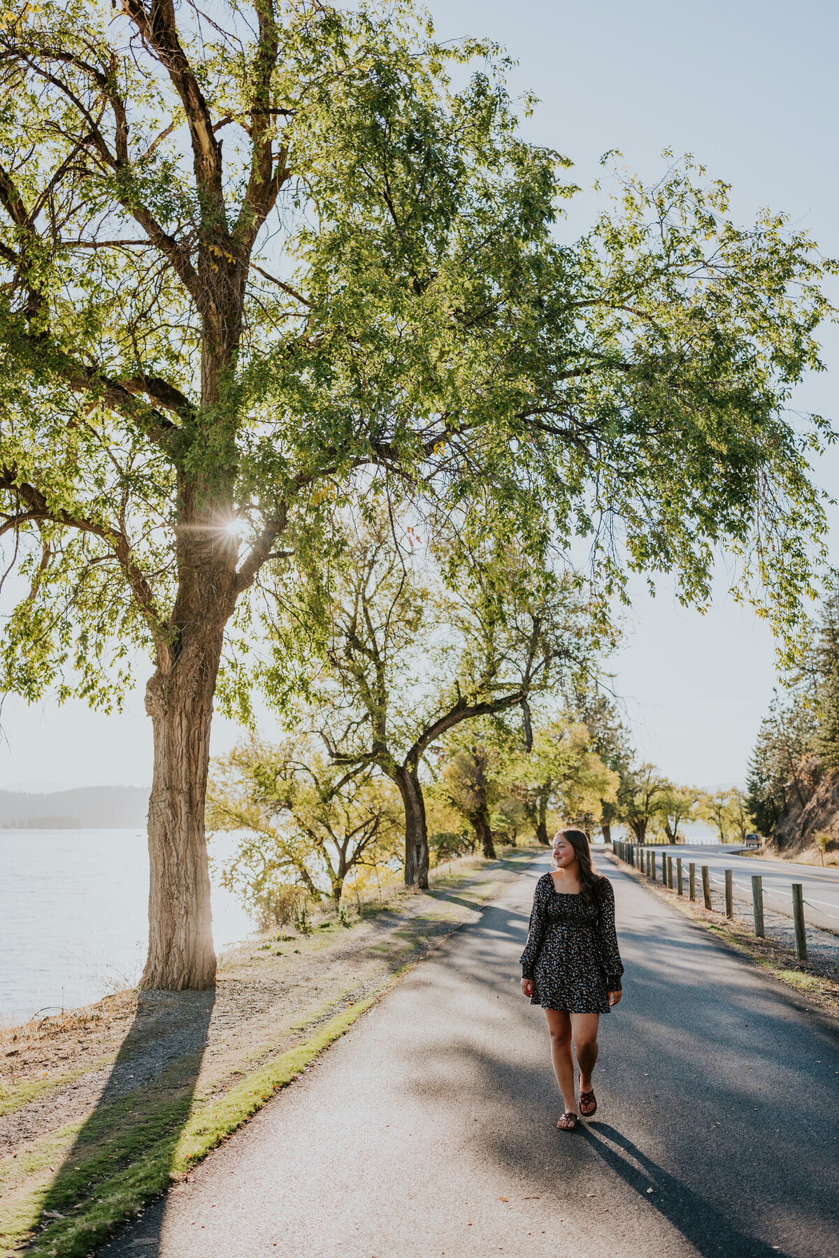 Young woman in cute dress walks along Centennial Trail  while sun shines behind tree.