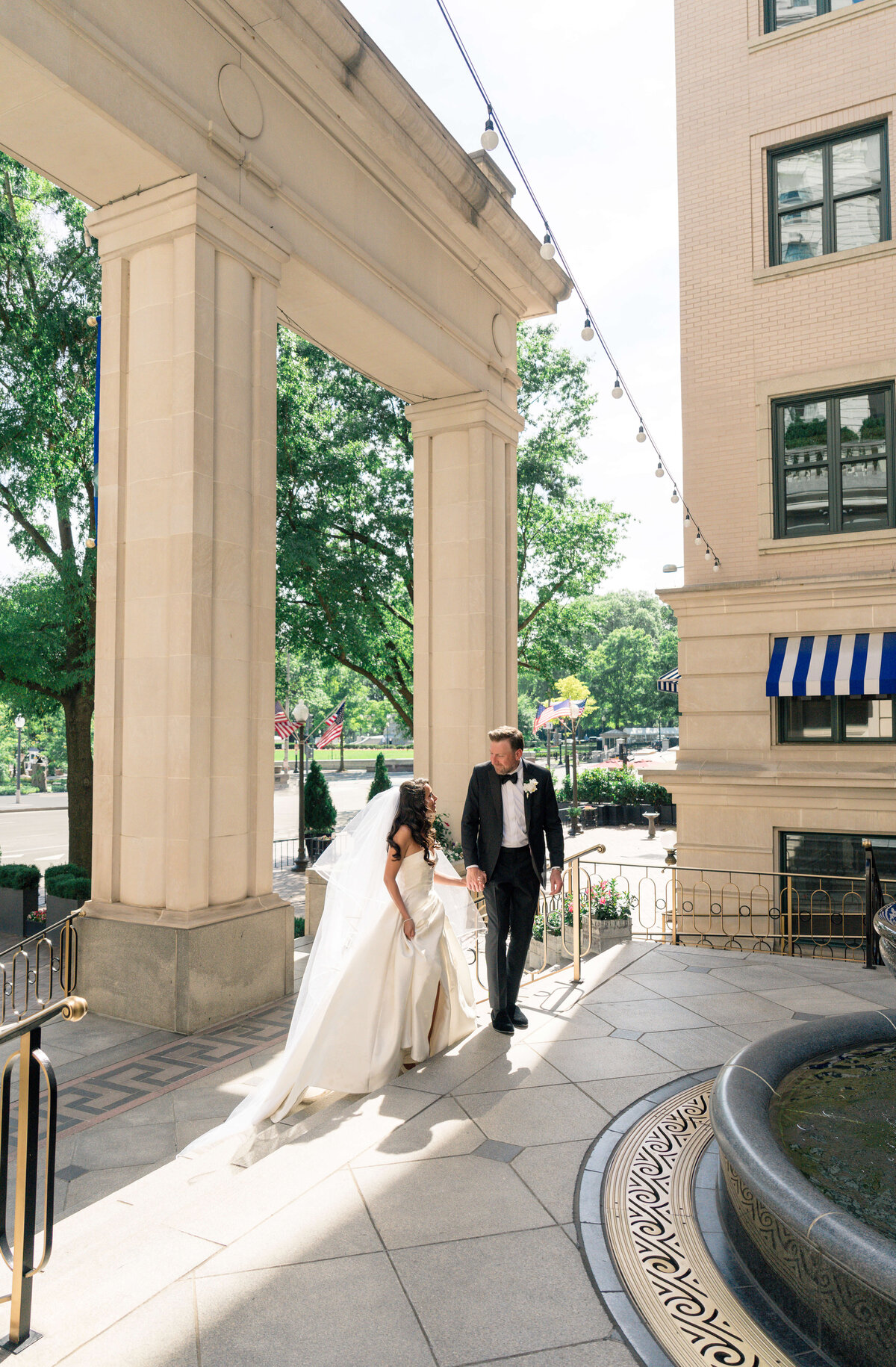 A bride and groom walk hand in hand under a large stone archway. The bride is wearing a white gown with a long veil, while the groom is in a black tuxedo. They are surrounded by tall buildings and greenery on a sunny day.