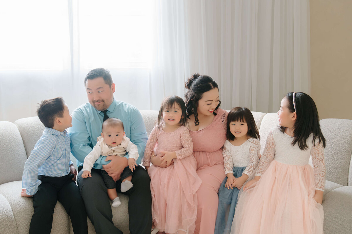 Family of seven sitting on a cream couch, smiling and relaxed under soft natural window light in the studio.