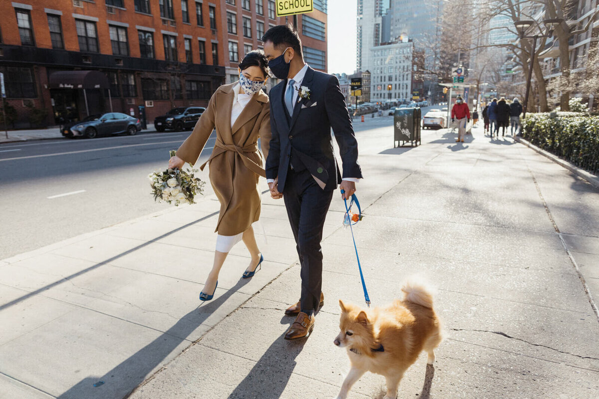 The bride, wearing a brown coat on top of her wedding gown, the groom, and their dog are walking in the streets of Brooklyn.