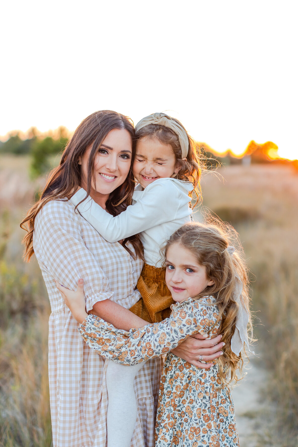 A joyful mother holds her two daughters close in a golden Virginia field at sunset, their laughter and love captured in a glowing moment by Brooke Tucker Photograph.
