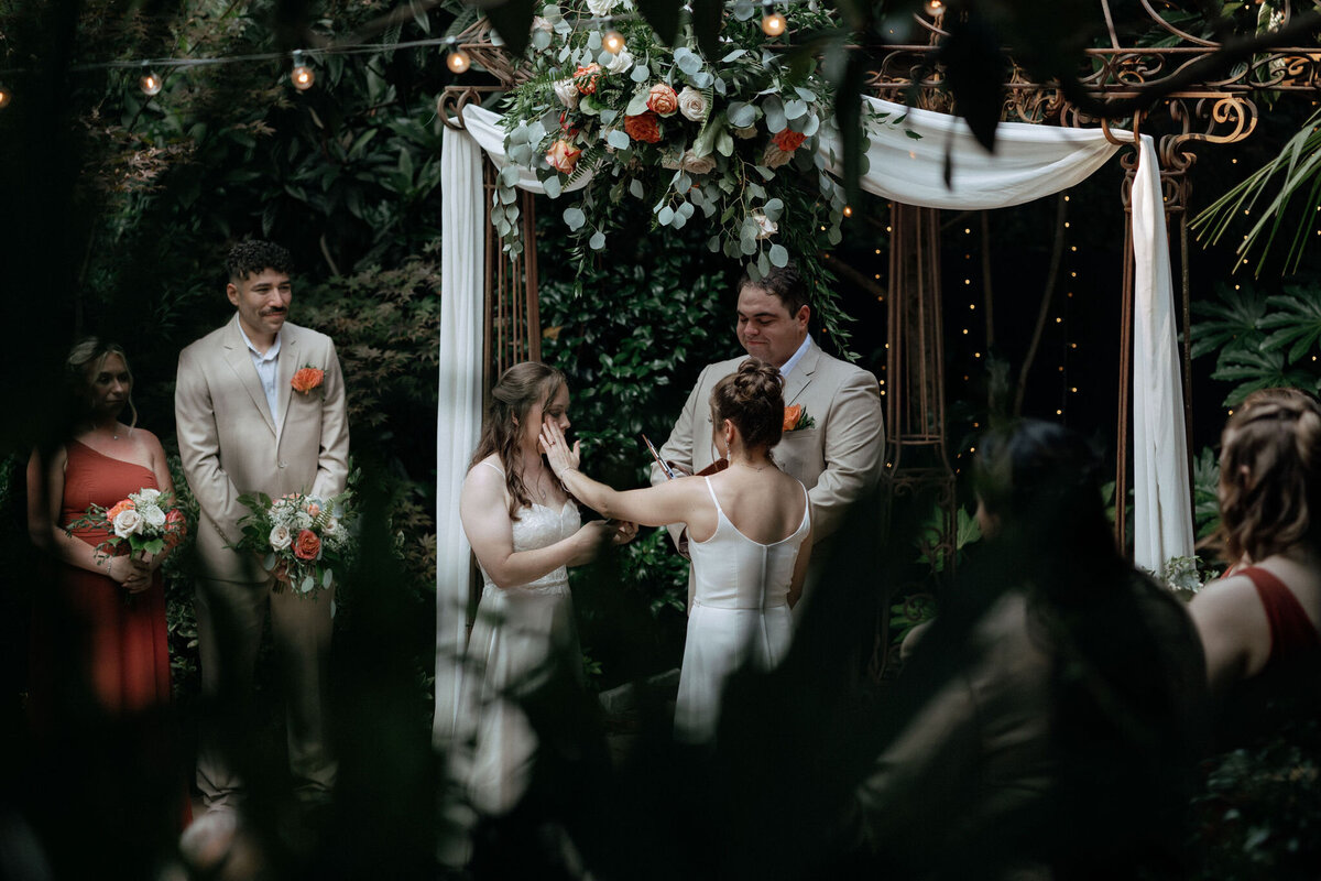 Two brides stand under a steel wedding arch as they recite their vows to each other. One bride wipes a tear away from the other bride's face.
