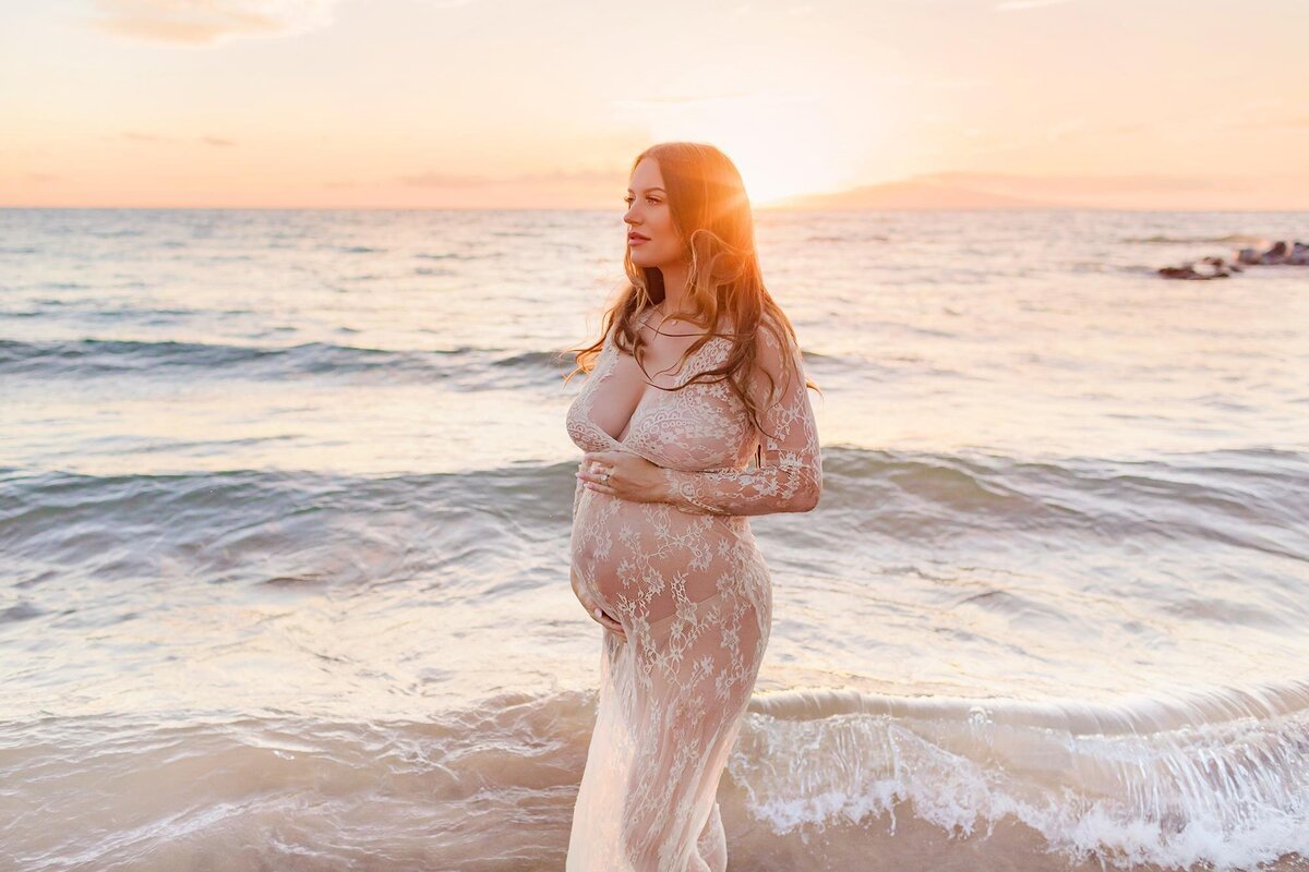 Stunning brunette wearing a white lace maternity gown poses next to the ocean at sunset on Maui