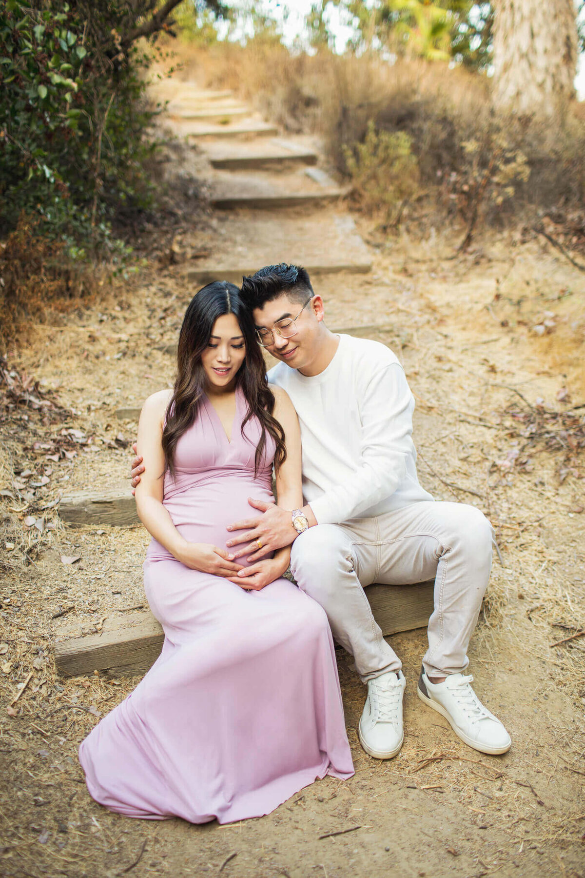 A maternity portrait of a couple sitting together on wooden steps in a natural outdoor setting. The pregnant woman wears a flowing lavender gown and rests her hands on her baby bump, while her partner, dressed in a white sweater and light-colored pants, lovingly embraces her from behind. He leans in with his head resting on hers, eyes closed, sharing a tender moment of connection and anticipation. The earthy surroundings, with dry leaves and a dirt path, create a serene backdrop that complements the couple's peaceful and intimate pose.