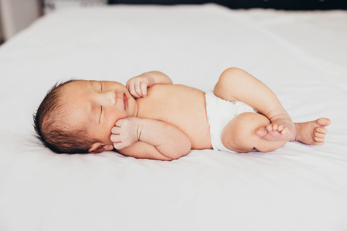 a sweet baby in a white diaper lays on a bed during his lifestyle newborn photography session in Encinitas