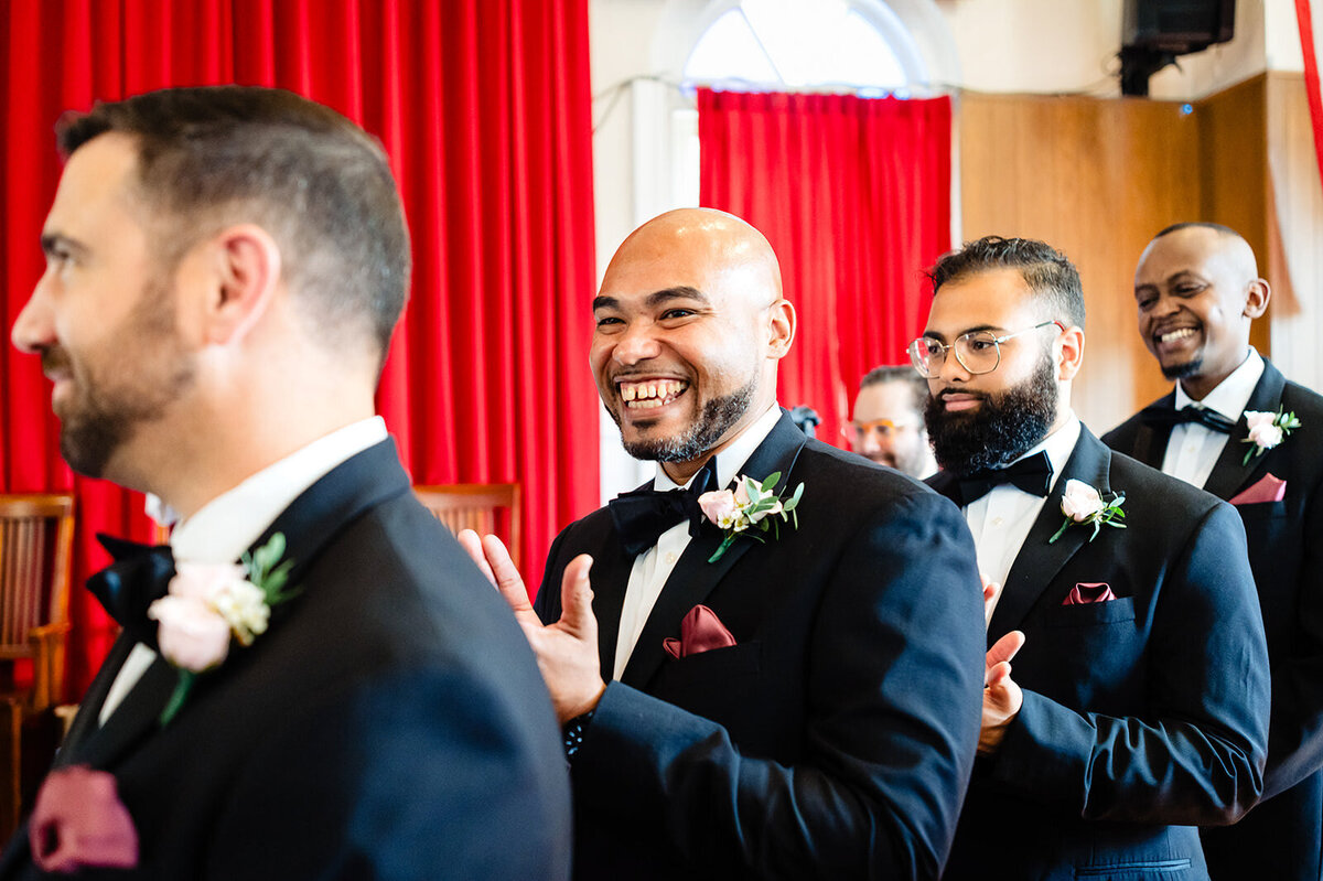 Groomsmen in black tuxedos with burgundy accents share a laugh, creating a lively atmosphere during a wedding ceremony