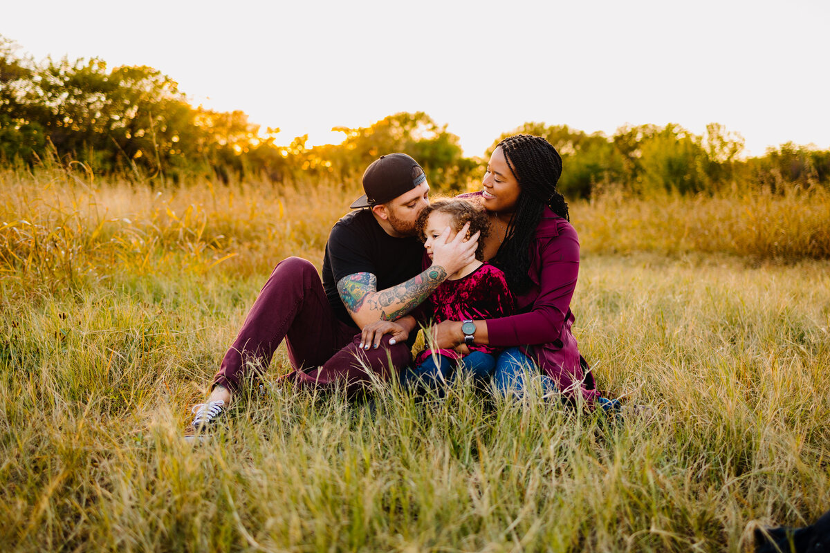 A touching moment captured in Albuquerque where the father is kissing his son's head with love, while the mom looks on with an adorable expression. The family is dressed in coordinated red and blue outfits, showcasing their warmth and affection