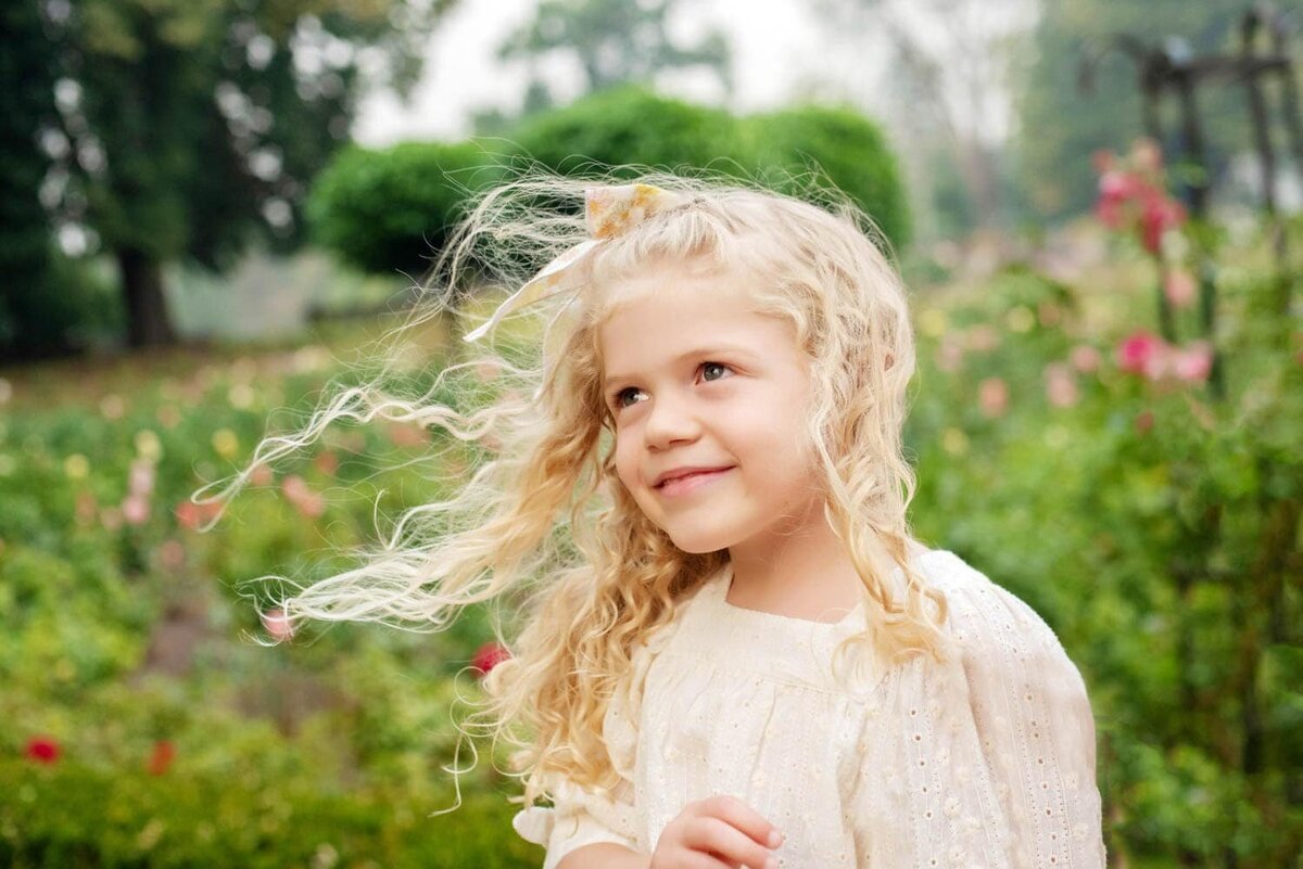 Little girl standing in rose garden with curly blond hair blowing in the wind.