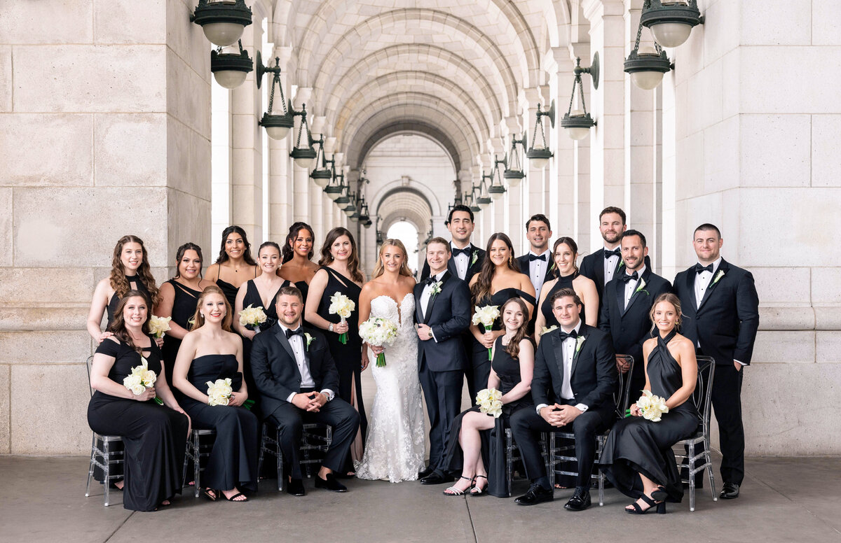 A bridal party smiles and poses in a grand stone archway. The bride and groom stand in the center, surrounded by bridesmaids in black dresses and groomsmen in black suits.
