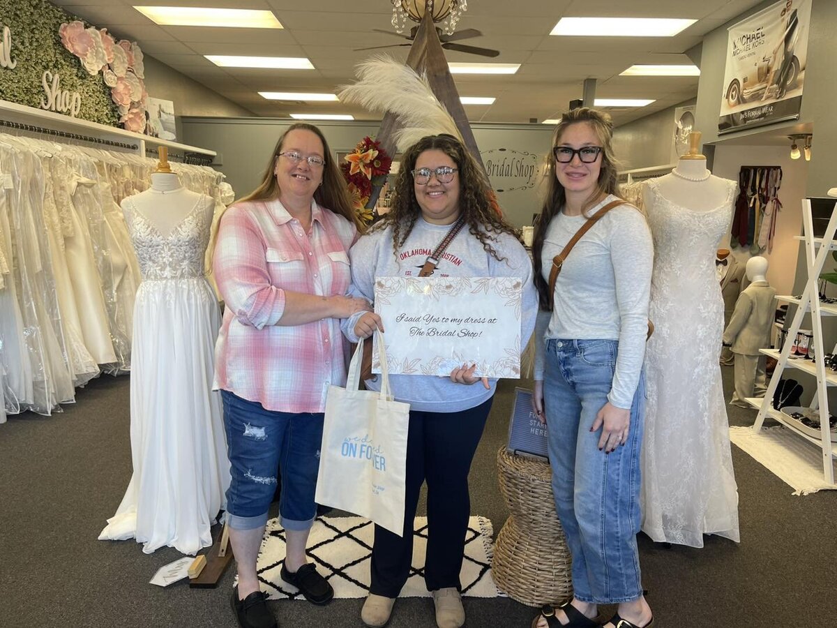 Three women standing together in a bridal shop holding a sign after choosing a dress