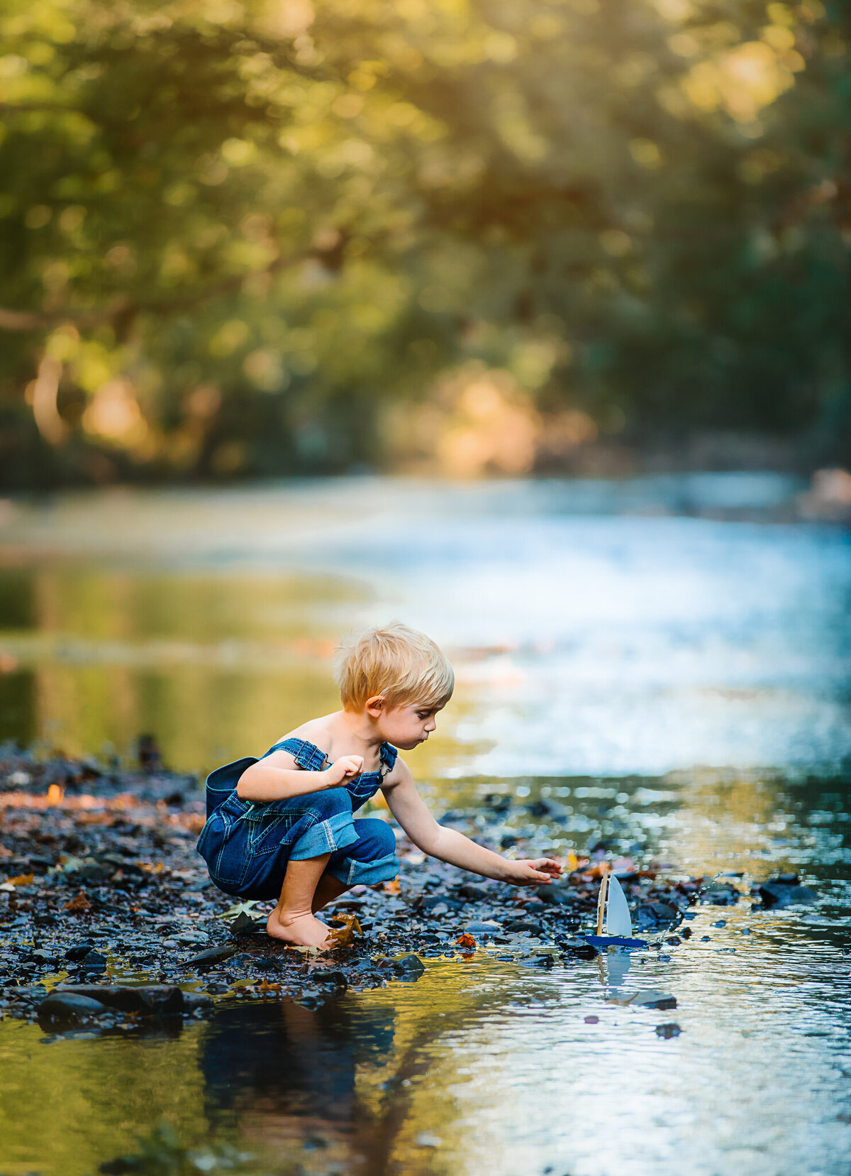 young boy in denim overalls kneeling down  to put toy boat in the river