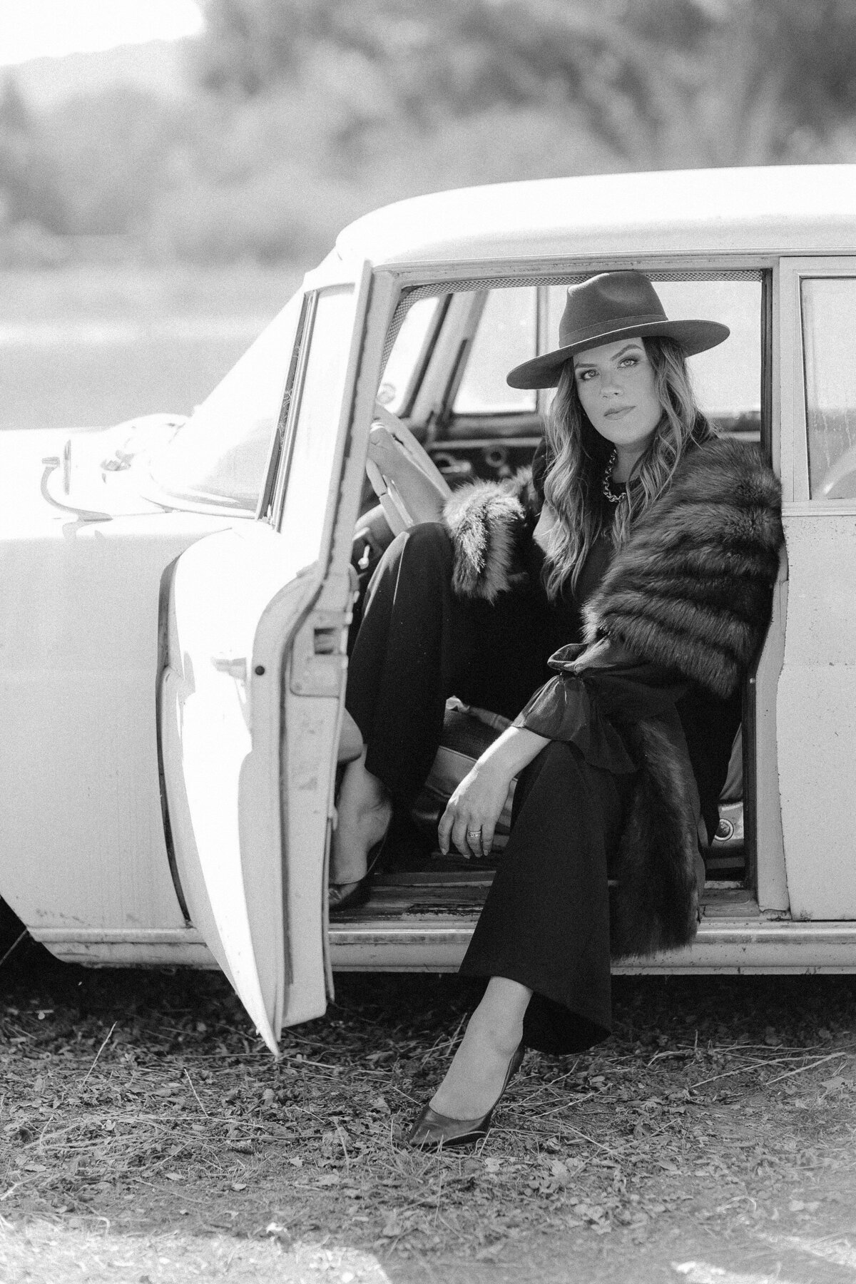 Wedding planner, Jessica Kuipers, poses for photo wearing hat and fur coat and sitting in drivers seat of vintage car