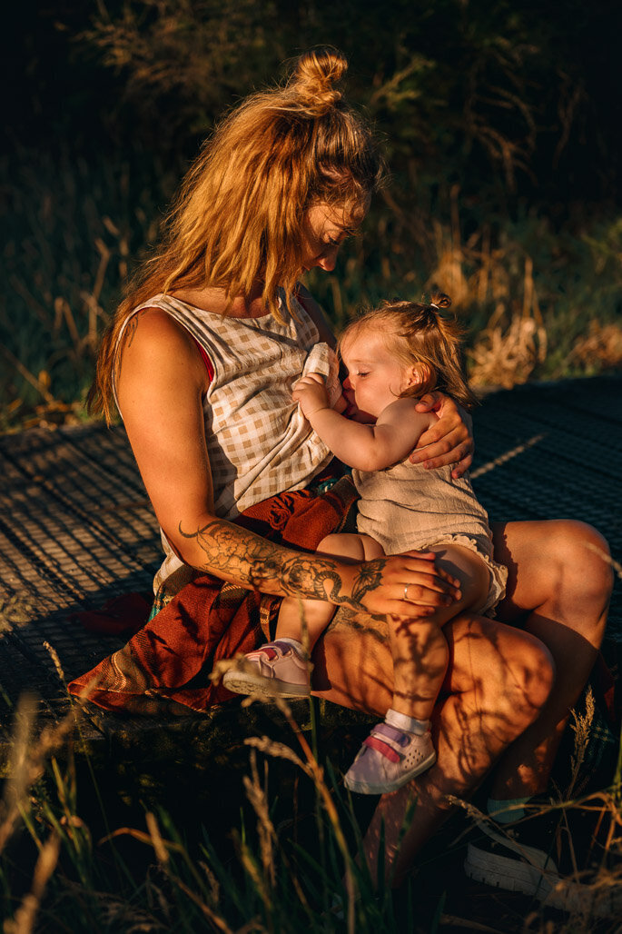 mum breastfeeding little toddler with the golden sun on them  and shadows of long grasses and mum is hevily tatooed