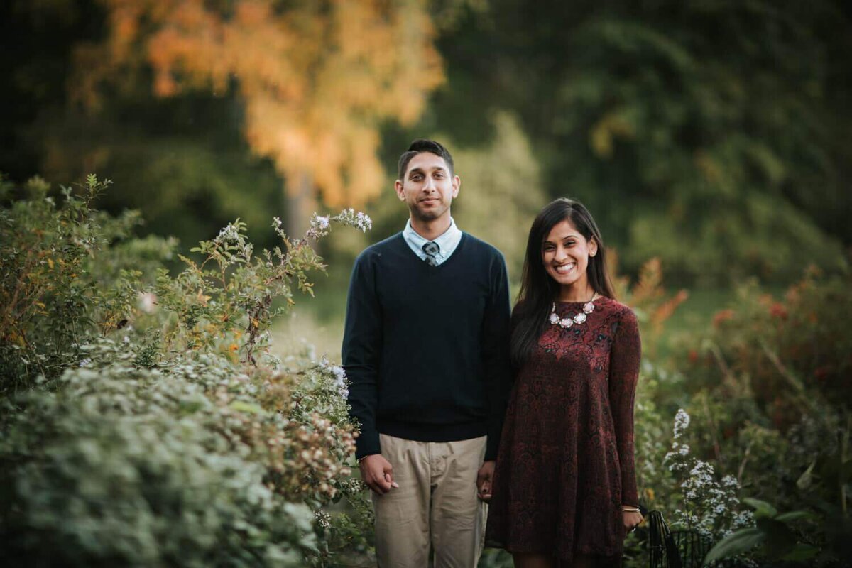 Couple standing side by side smiling and posing surrounded by greenery in New York.