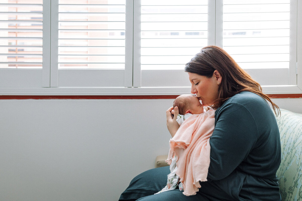 mom in green robe holding and kissing forehead of newborn wrapped in pink blanket