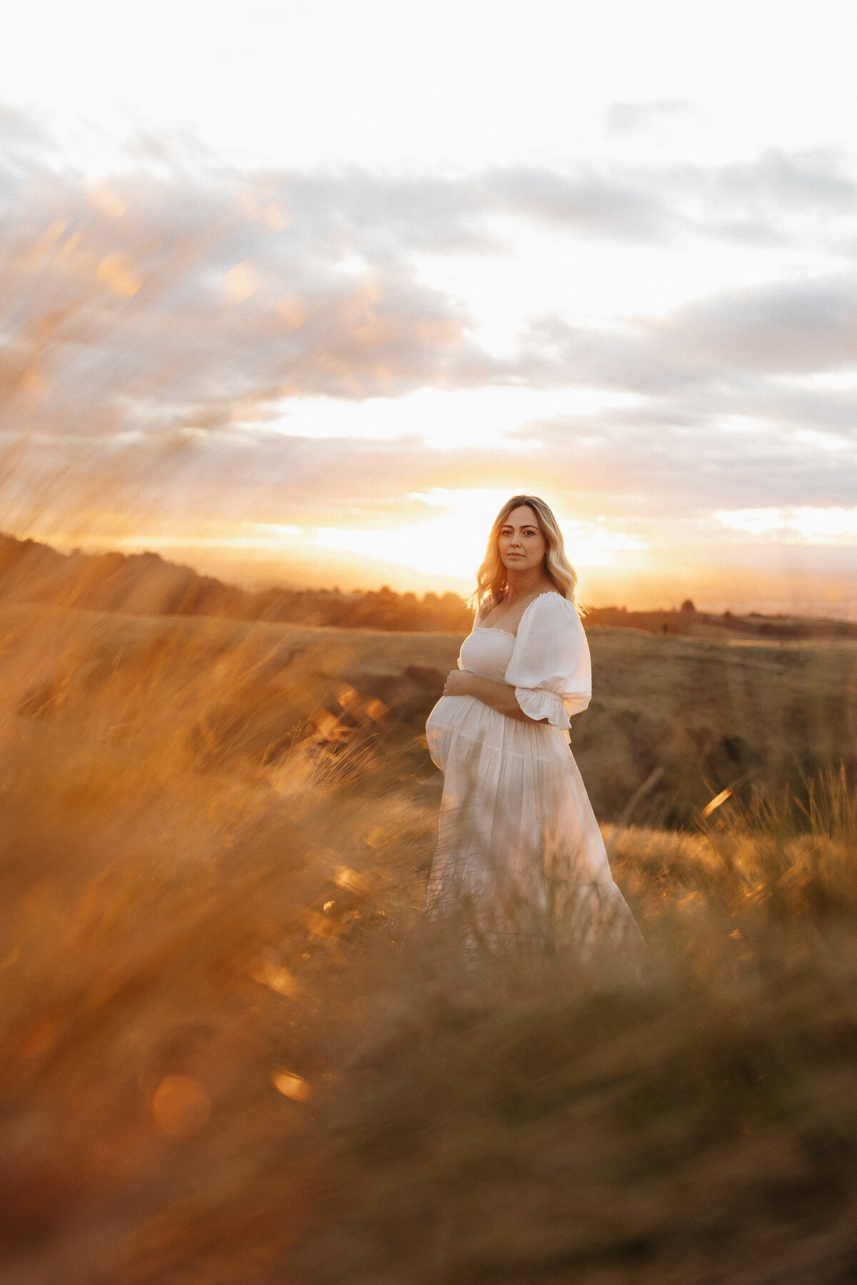 An expectant mother wears a beautiful white dress with puff sleeves during a pregnancy photoshoot in the hills surrounding christchurch at stunset with lucy jane photography