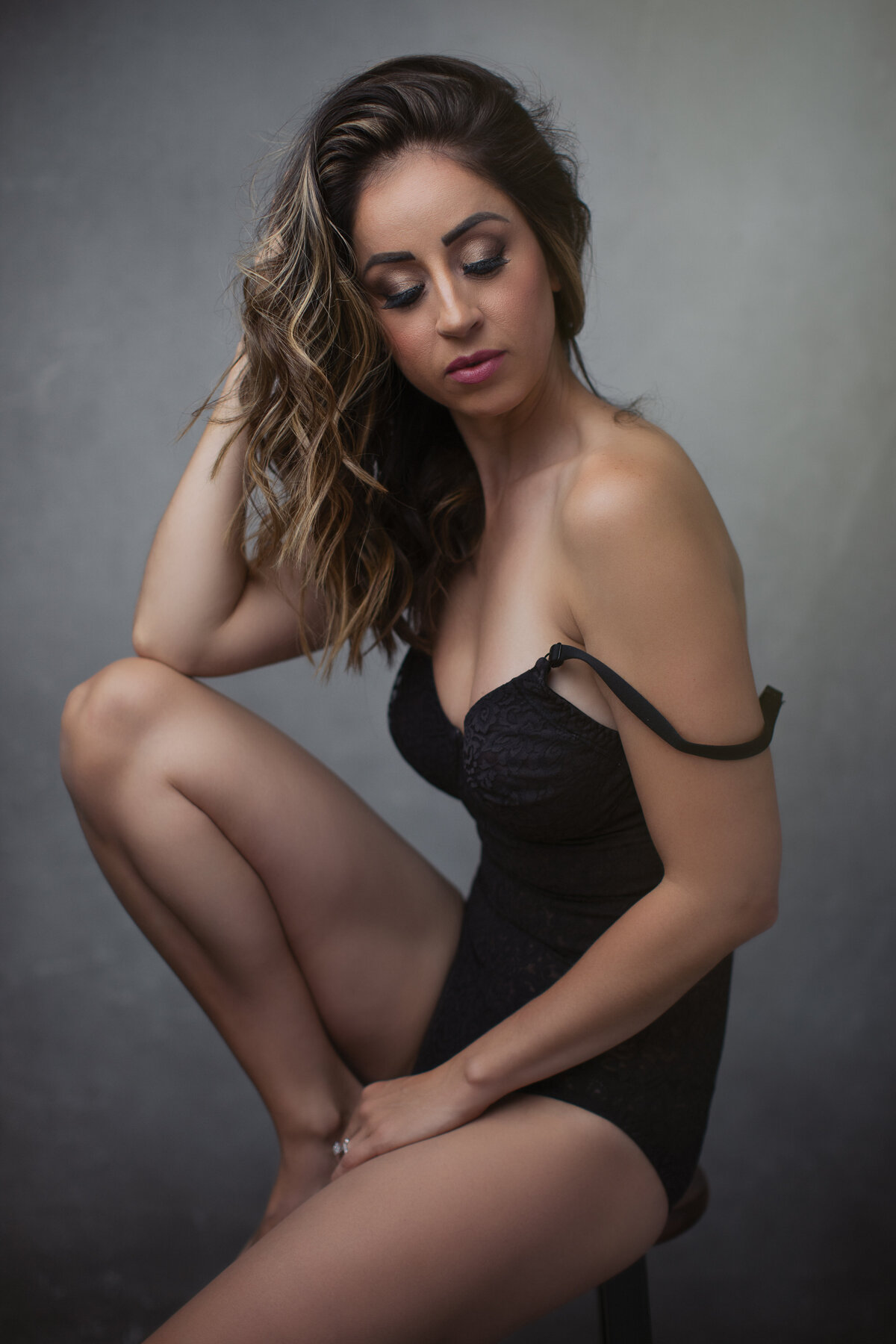 A woman wearing a black swimsuit sitting on a stool touching her hair and looking down