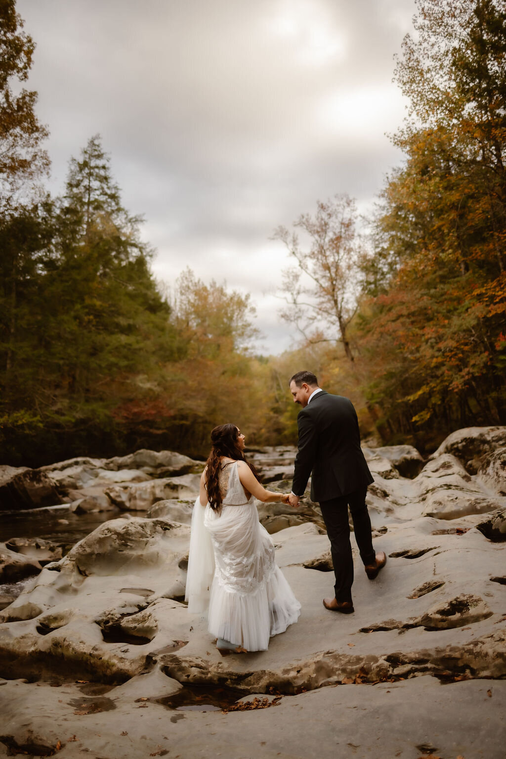 Bride and groom walking hand-in-hand across the textured stone riverbed at Greenbrier during their eloping to Gatlinburg, surrounded by moody skies and autumn-hued forest.
