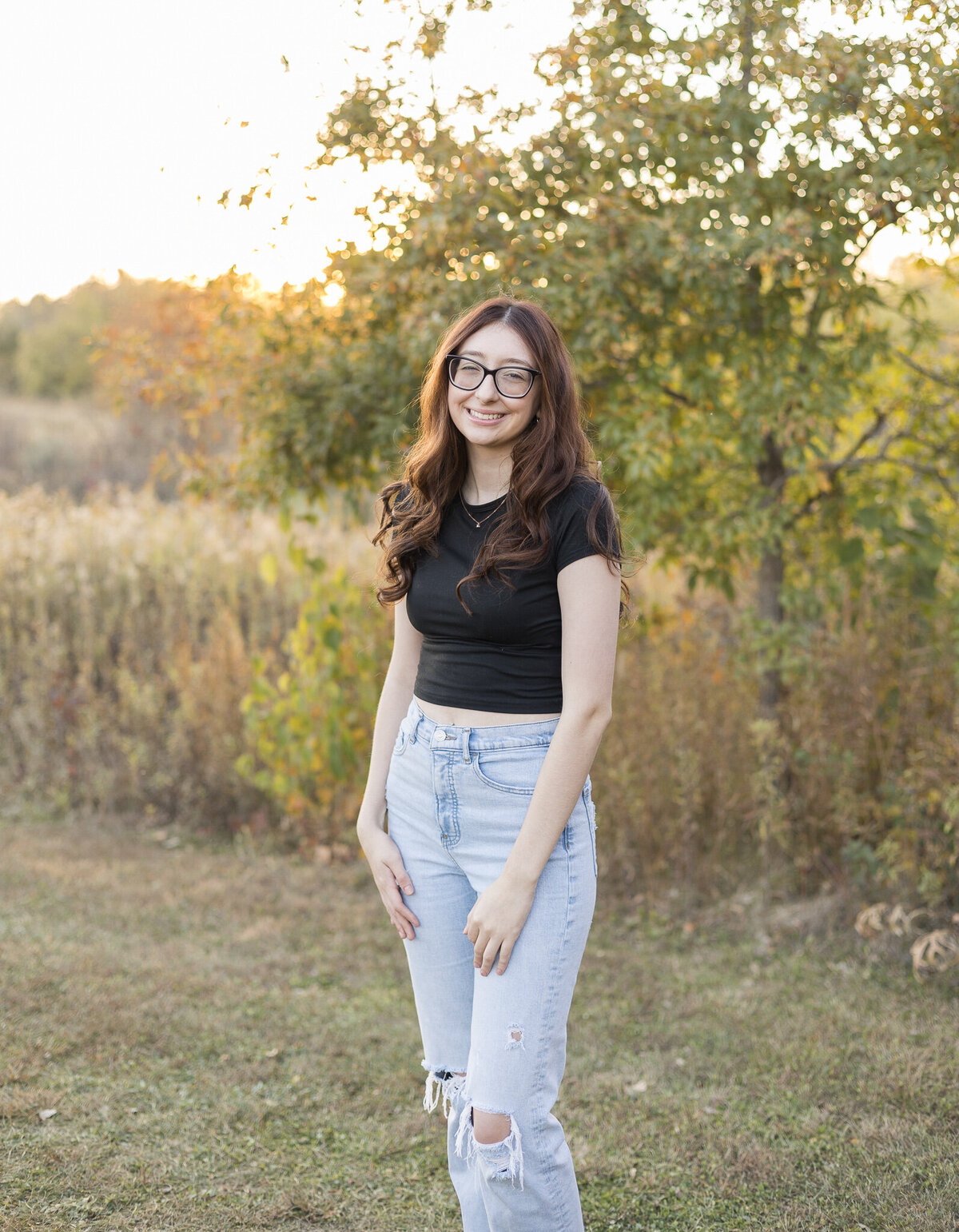 High school senior female, with long black hair, wearing glasses, blue jeans, short sleeve black shirt, standing in grass with trees in the background, smiling at the camera
