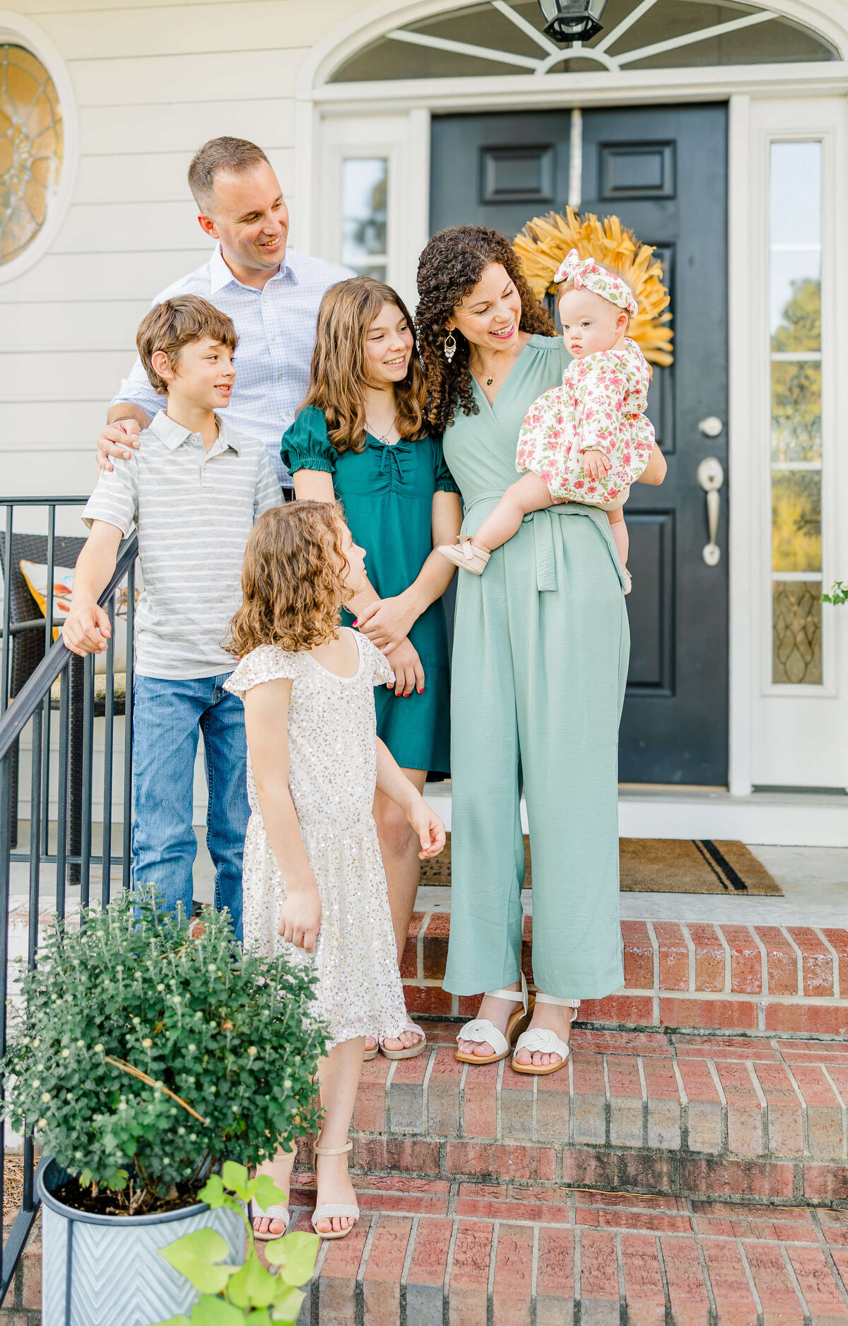 A Fayetteville NC family photographer captures a touching scene: a family of six on the steps of a house with a black door. The adults cradle their baby, while three children stand close. A potted plant and decorative glass accents add charm to this beautifully framed moment.