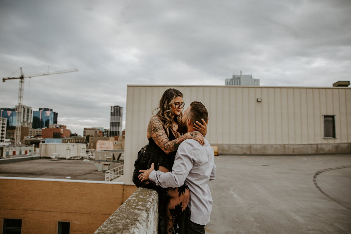 Downtown London, Ontario rooftop parking garage engagement session. Woman is sitting on the ledge with her husband between her legs. His hands are on his thighs, her hands are wrapped around his neck. They are looking at each other in this intimate photo.